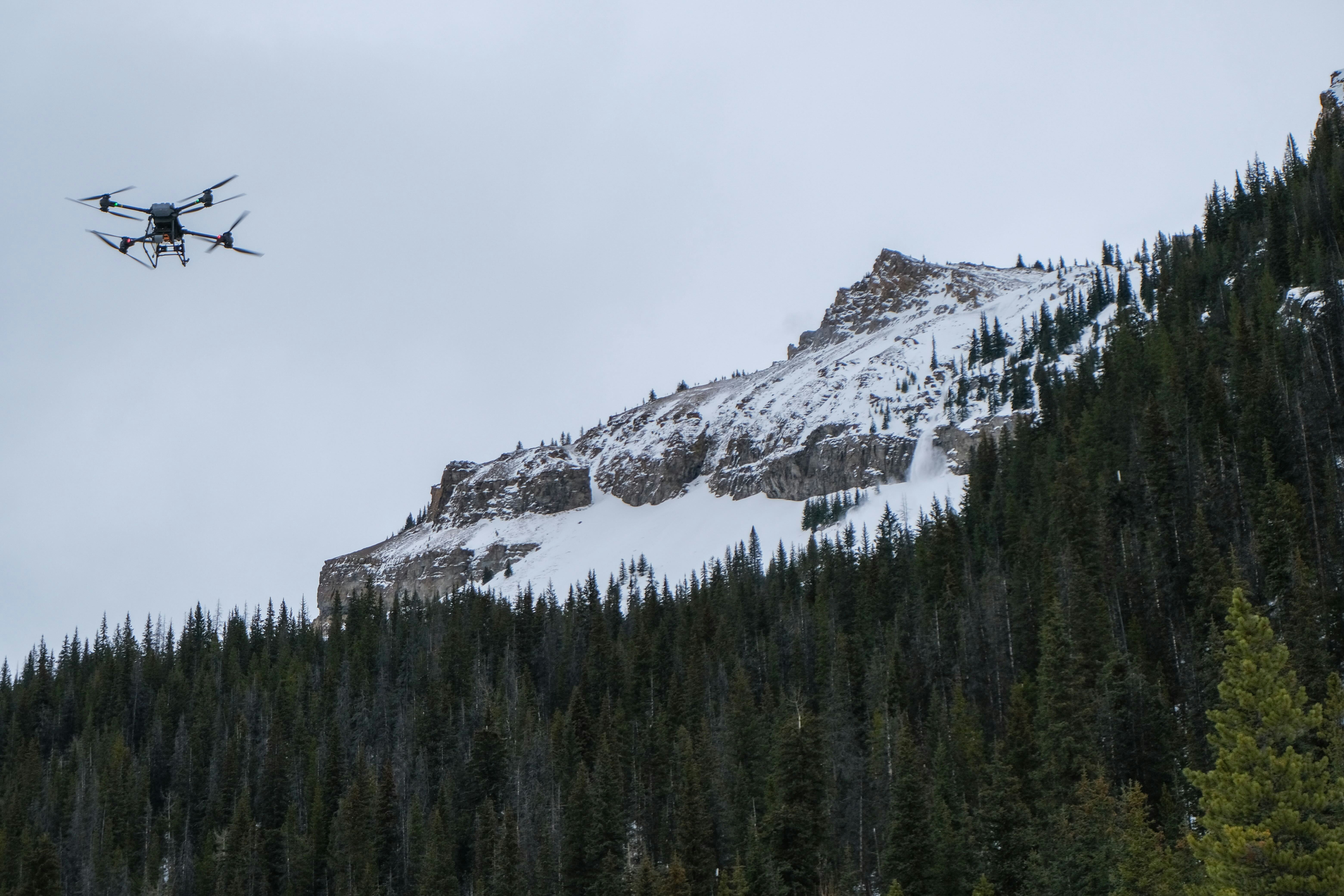 A drone flies over snow-covered mountains and evergreen trees.