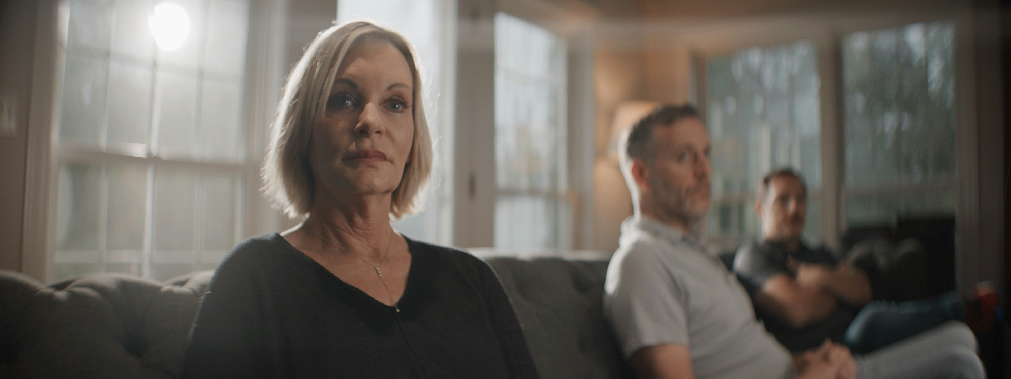 A blonde woman in her fifties sits on a grey sofa, looking toward the camera with a steady expression while two men talk quietly behind her in a sunlit room.