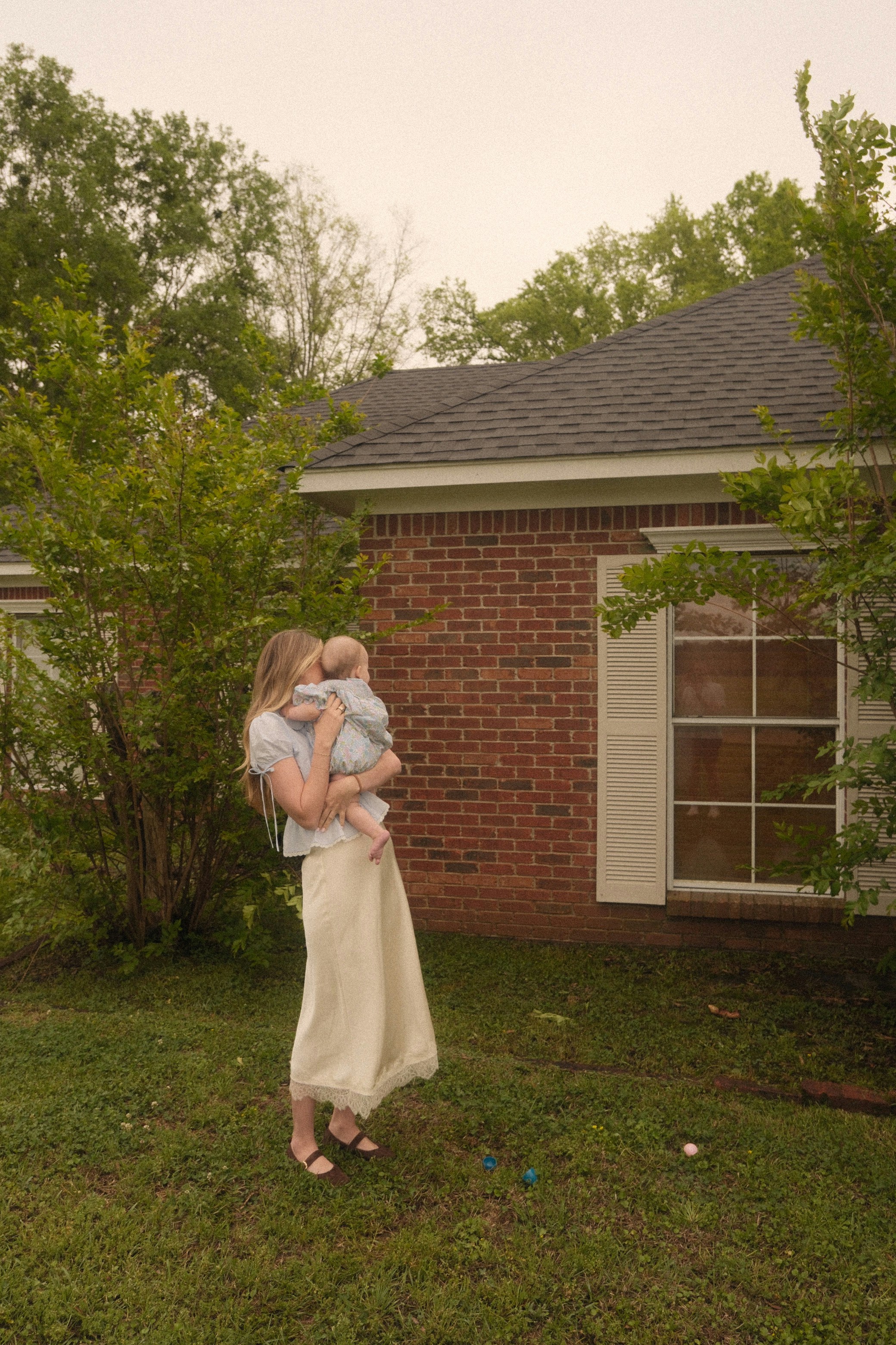 Mother holding baby outdoors near a brick house.