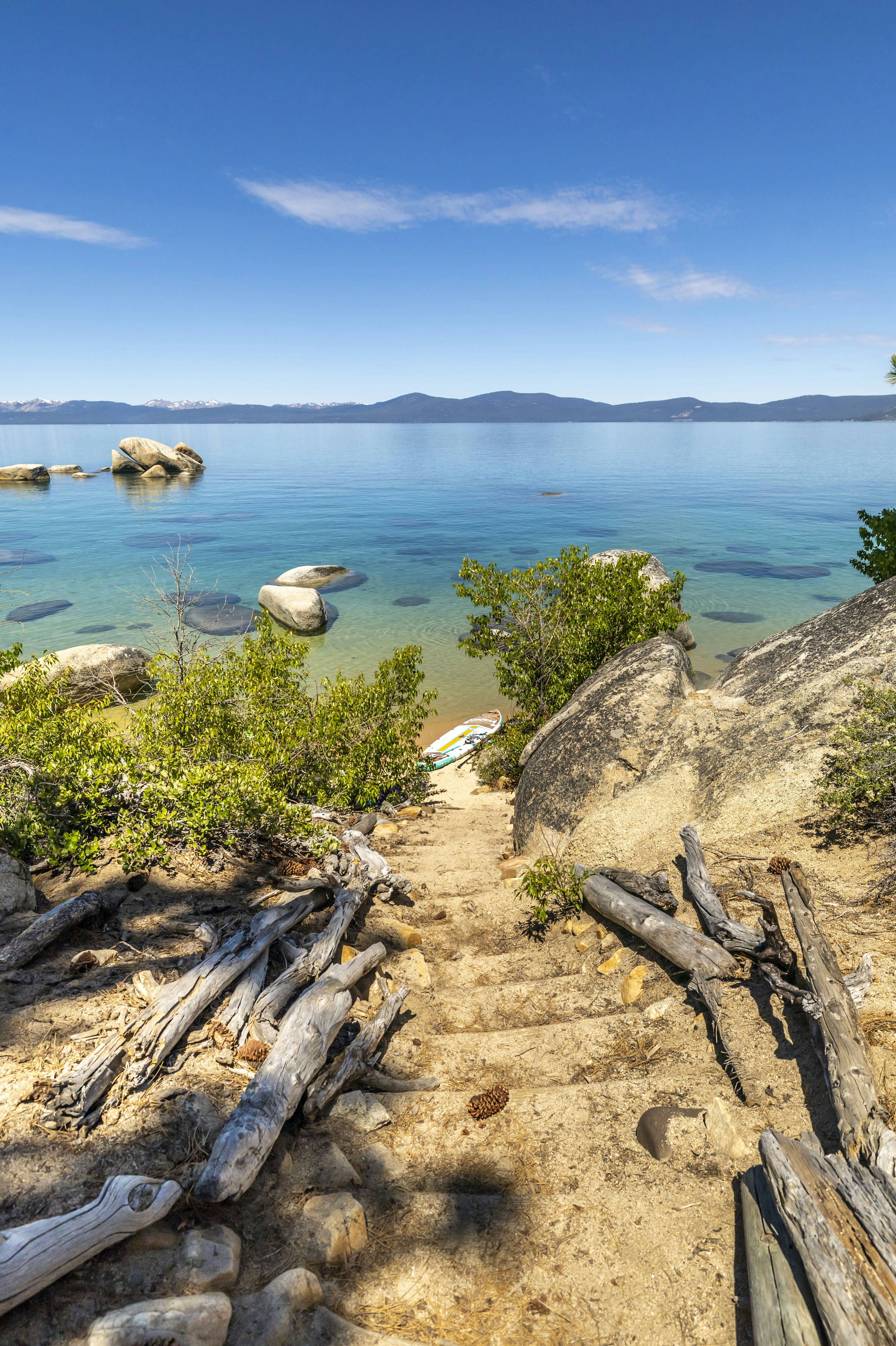 Clear blue lake with sandy path, driftwood, and distant mountains.
