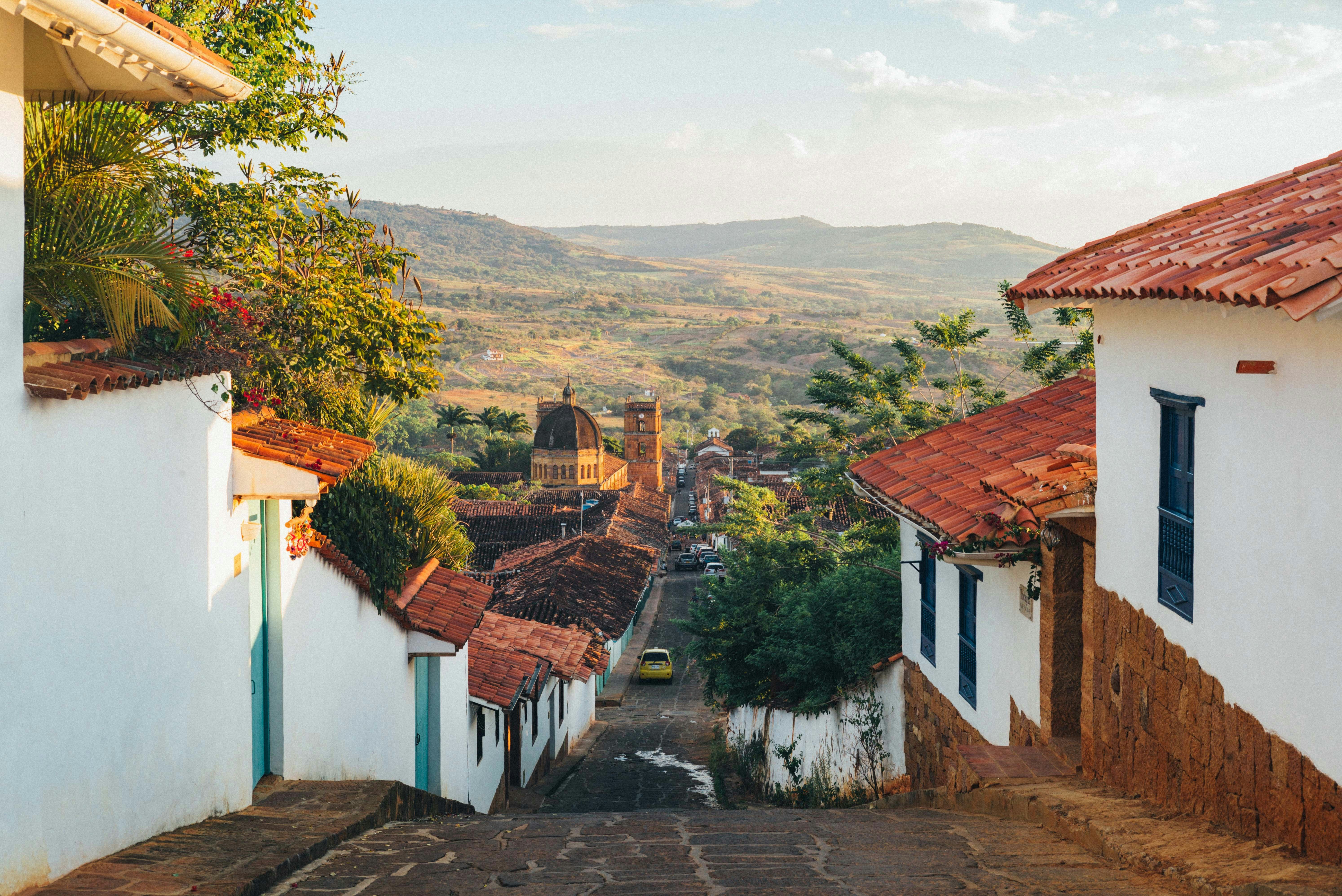Sunlit village street lined with white walls and red-tiled roofs recedes toward distant hills. A narrow stone path leads the eye through the charming, warm-colored scene.