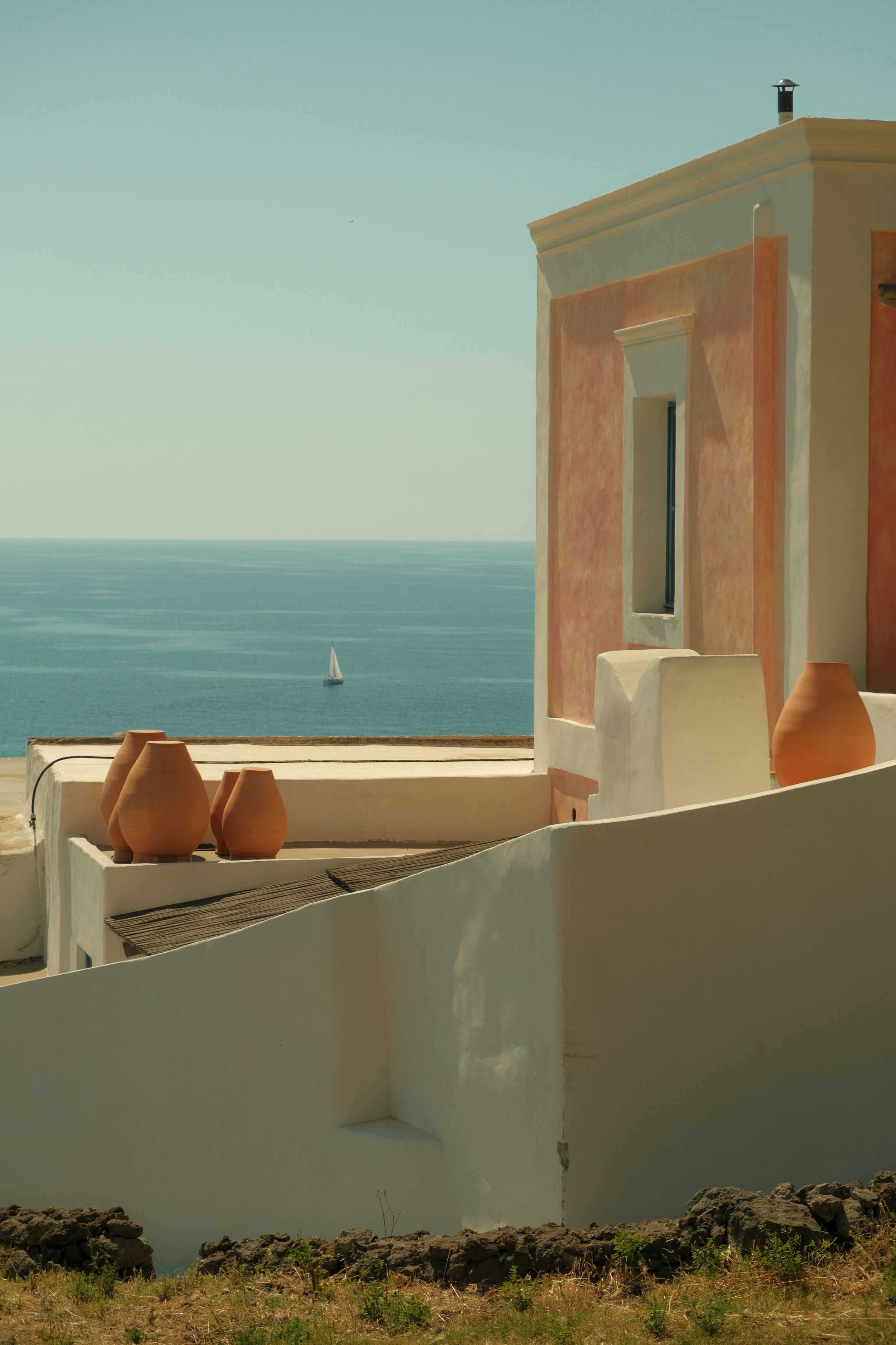 Sunlit coastal architecture with terracotta pots against a pale sky and sea horizon; the scene emphasizes warm stucco walls and white accents as a serene, Mediterranean detail.