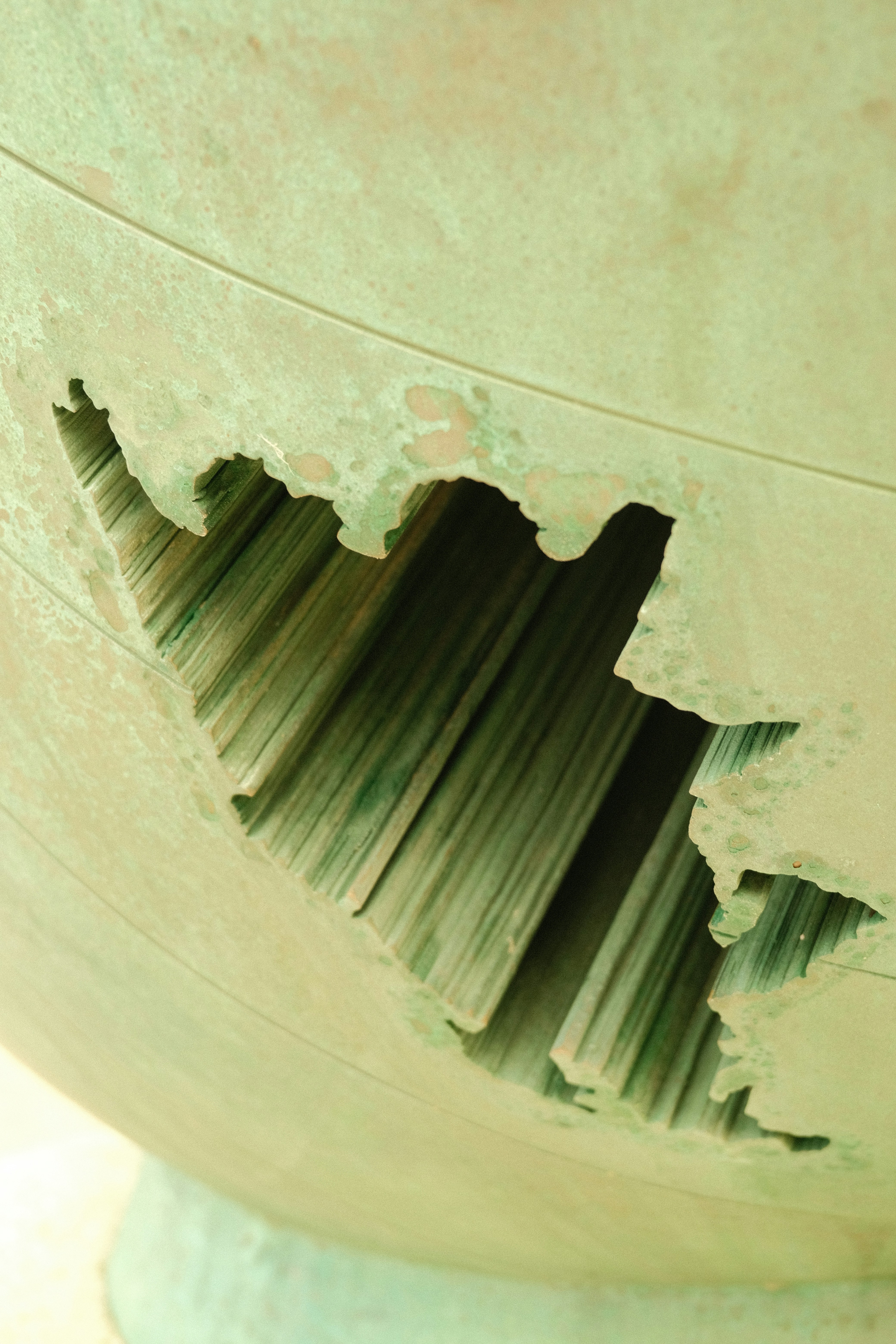 A close-up of a weathered, cracked ceramic surface revealing layered interior textures; the image emphasizes the jagged edge and striated interior with a pale green glaze. This is a photographed sculpture surface detail.