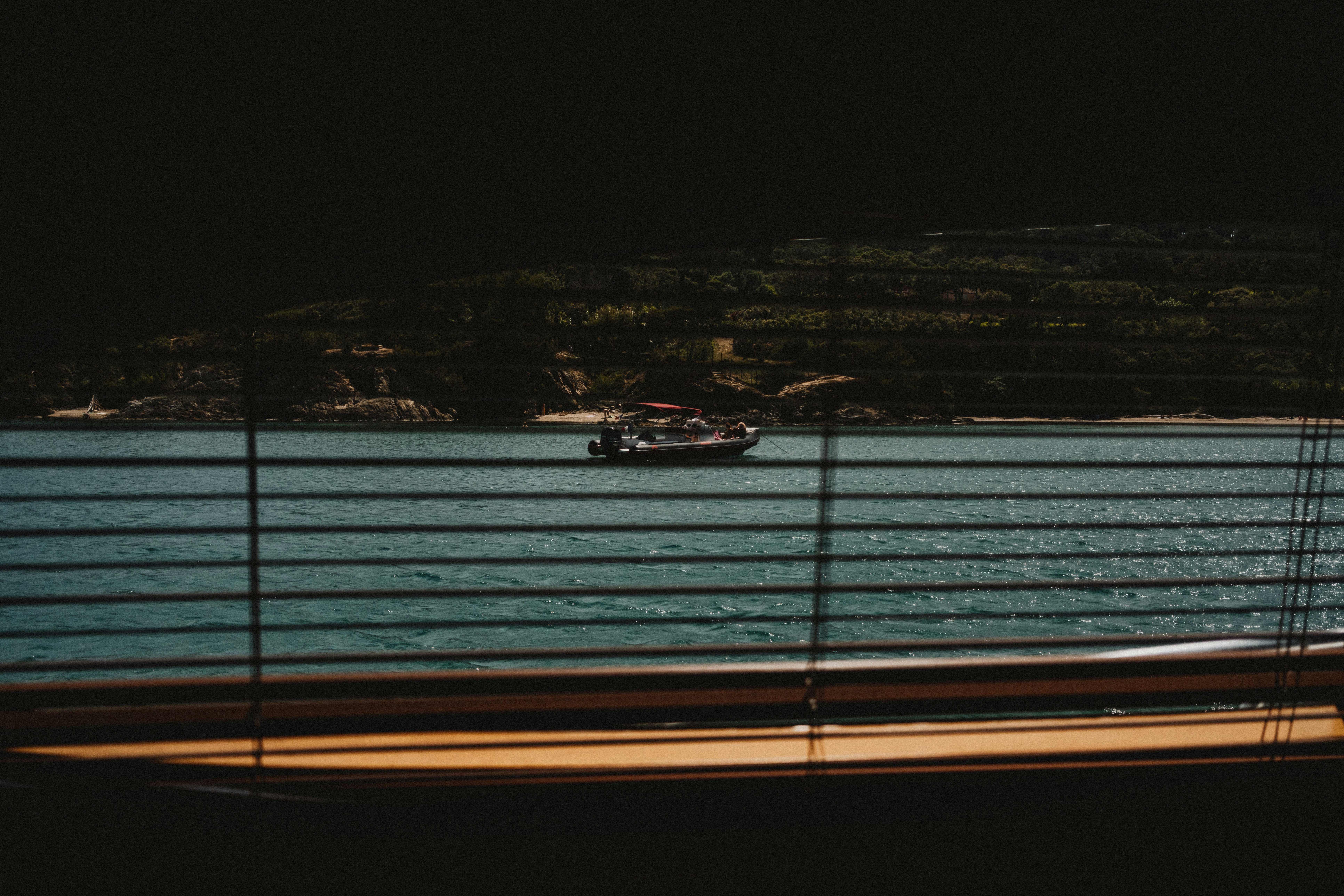 Boat on calm blue water, viewed through horizontal blinds.