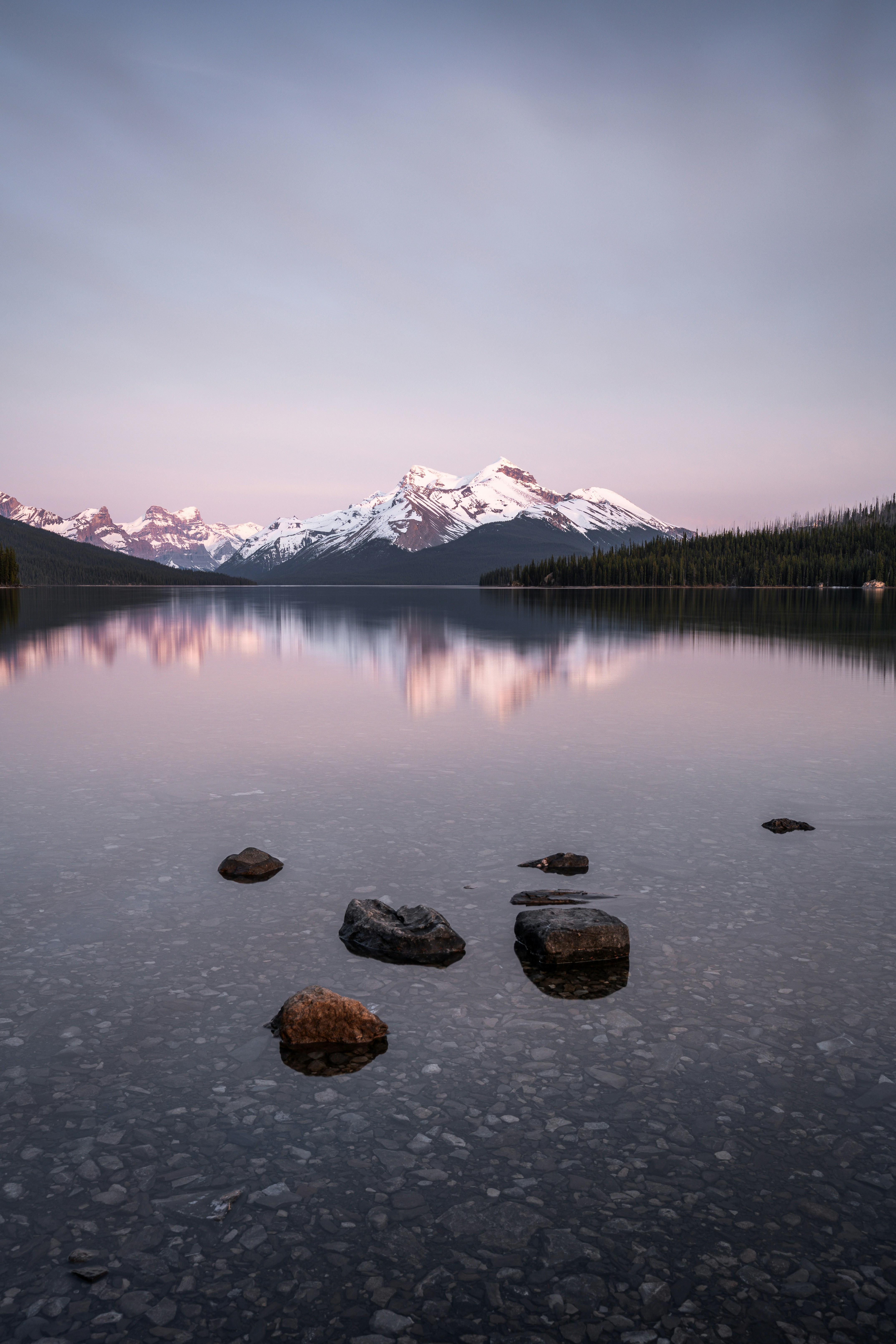 Schneebedeckte Berge spiegeln sich in einem ruhigen See mit sichtbaren Felsen.