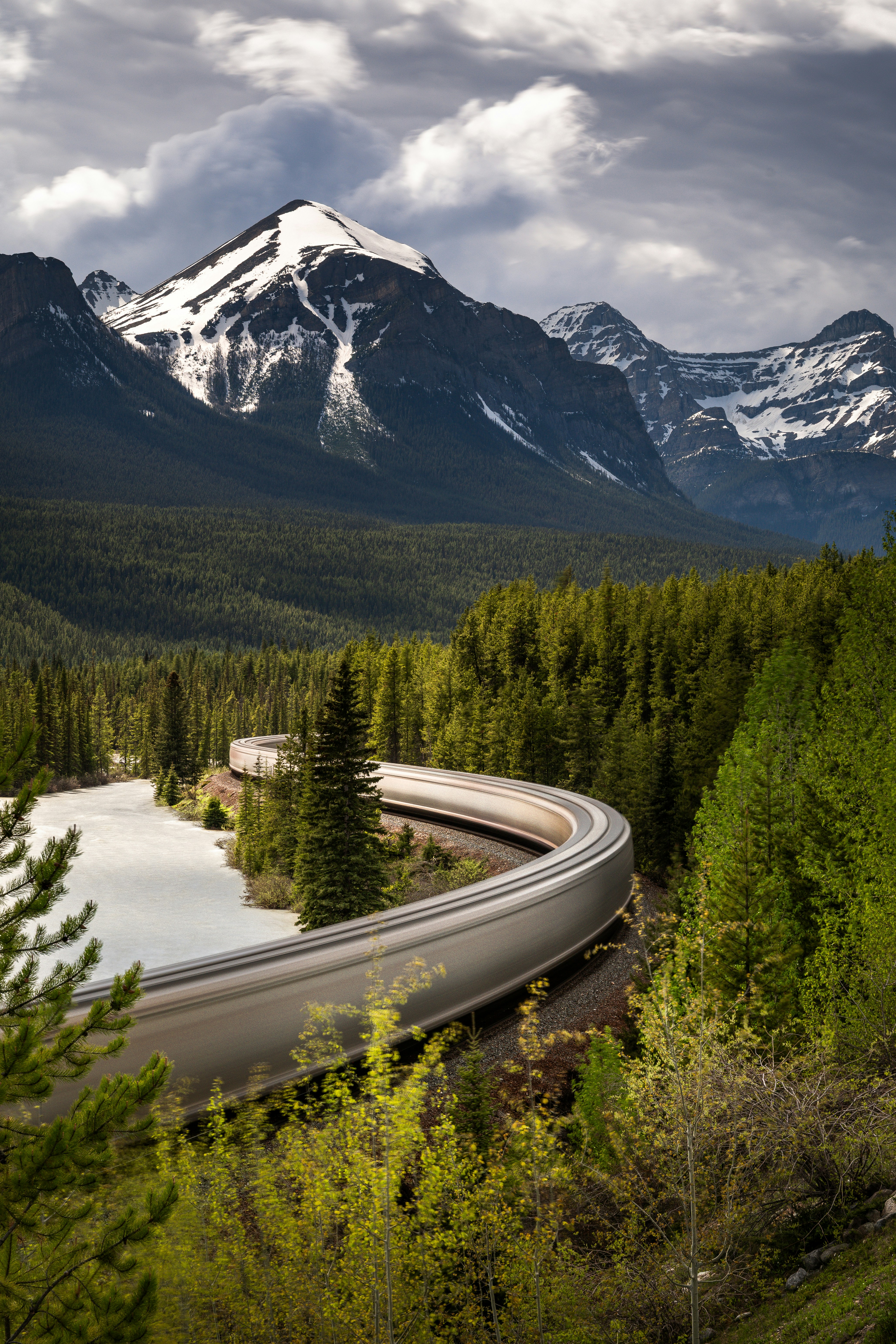 A train winds through a lush forest with snow-capped mountains.