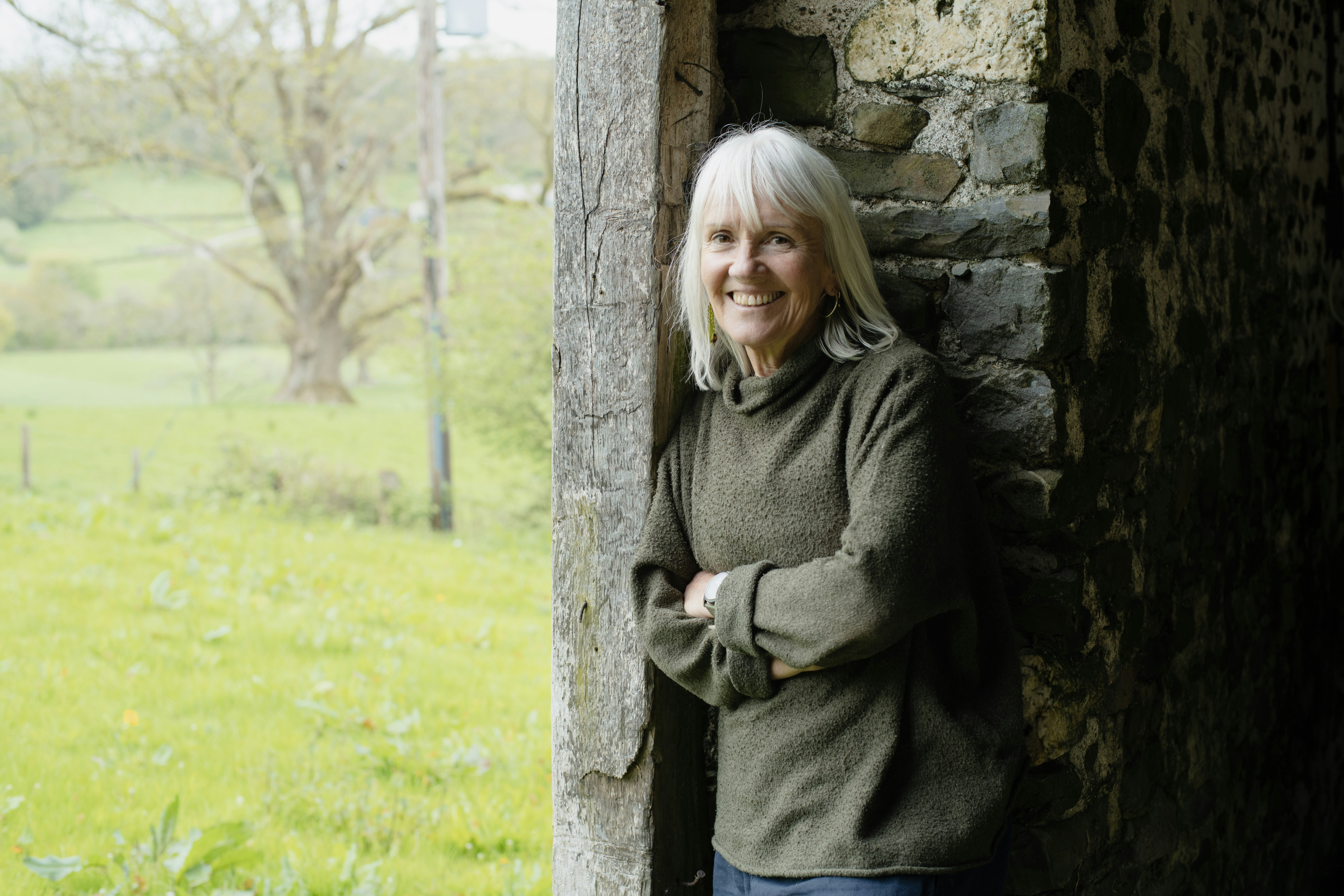 A smiling woman leans against a rustic stone wall outdoors.