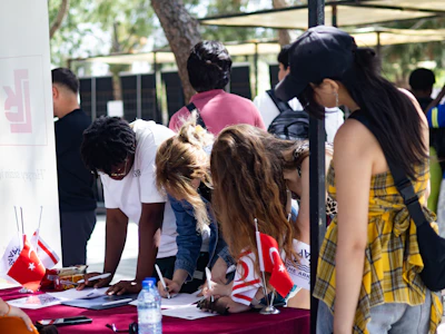 People signing papers at an outdoor table with flags.