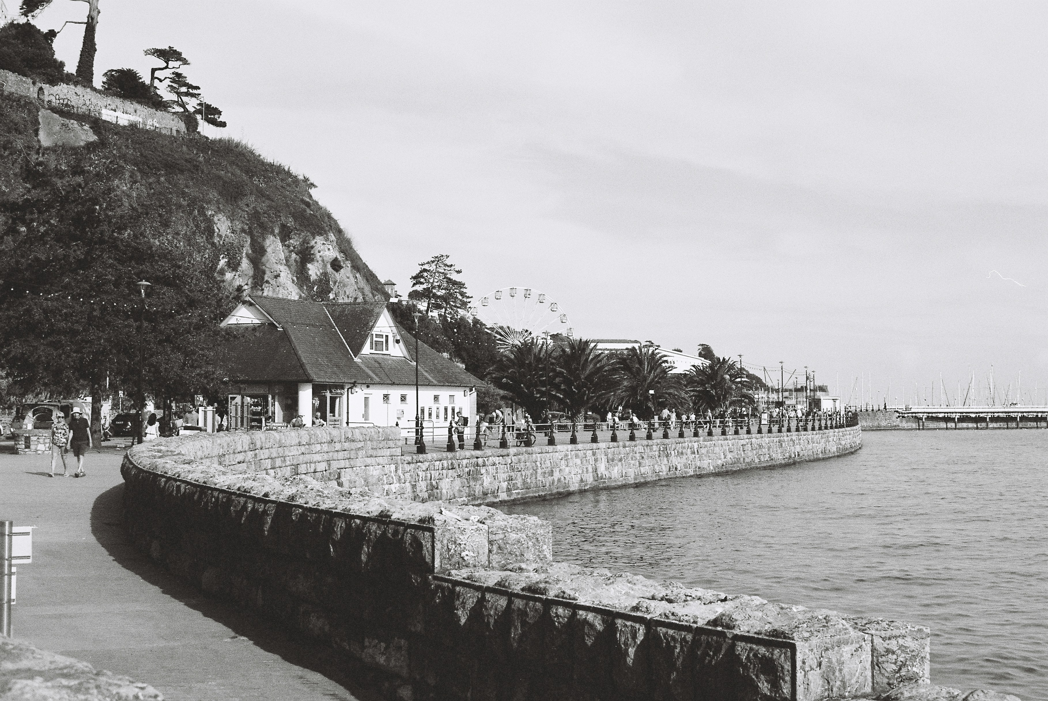 Paseo marítimo con muros de piedra, edificios y noria lejana.