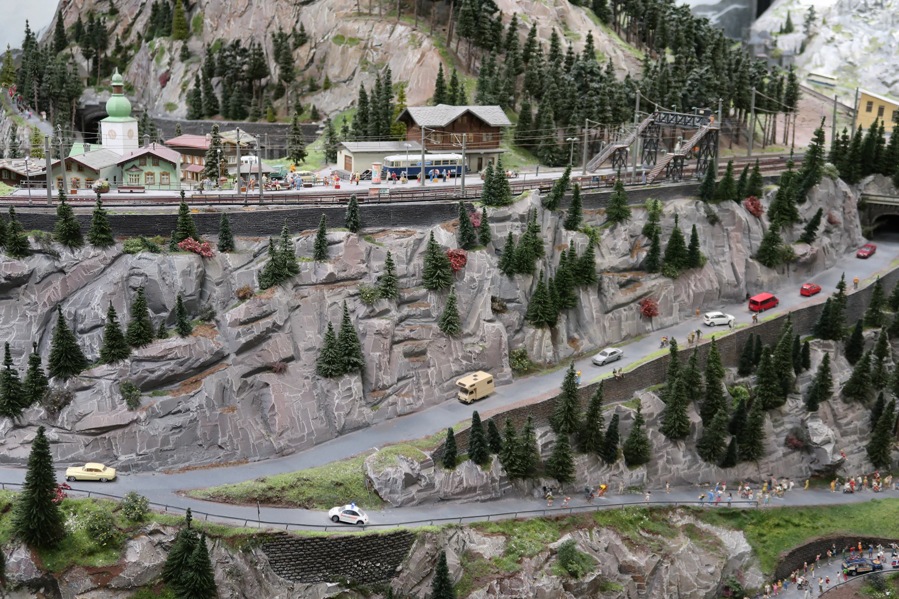 A large model mountainside of gray rock faces and dark green conifers, with roads and tiny cars winding across it