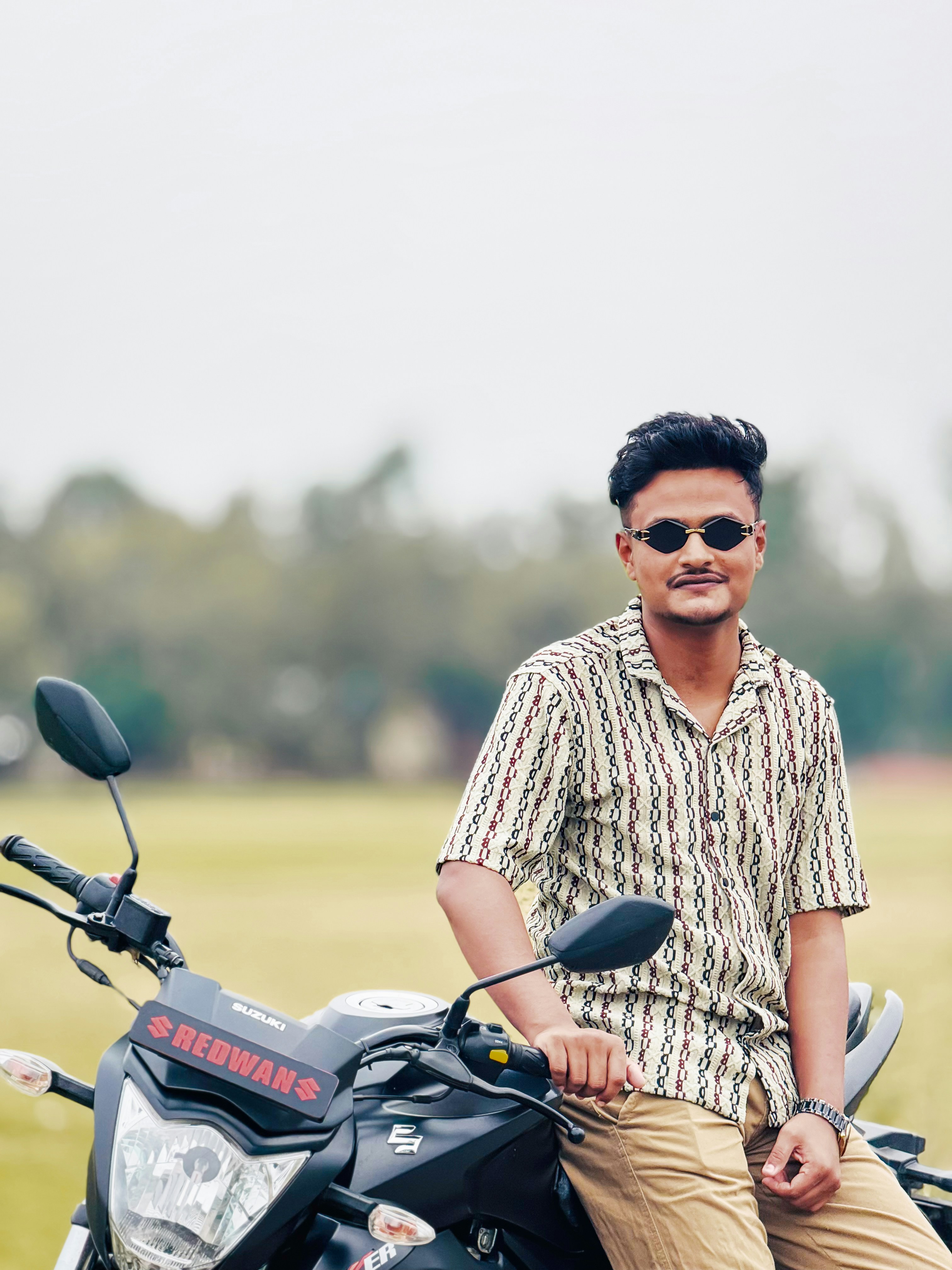 Pria muda bermotif kemeja dan kacamata hitam di atas sepeda motor.