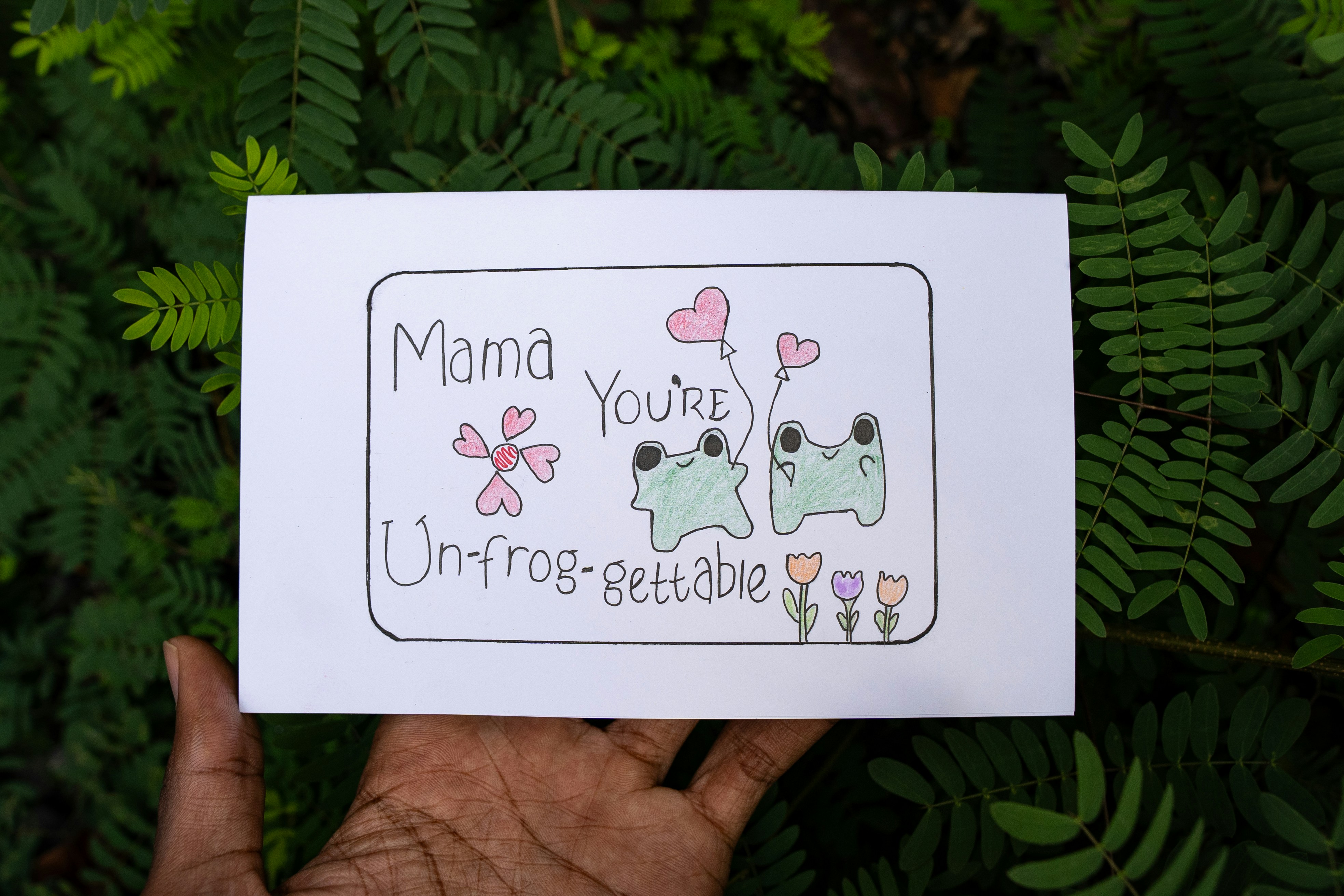 Segurando um cartão "mamãe, você é uni-sapo que pode conseguir" com desenho de sapo.