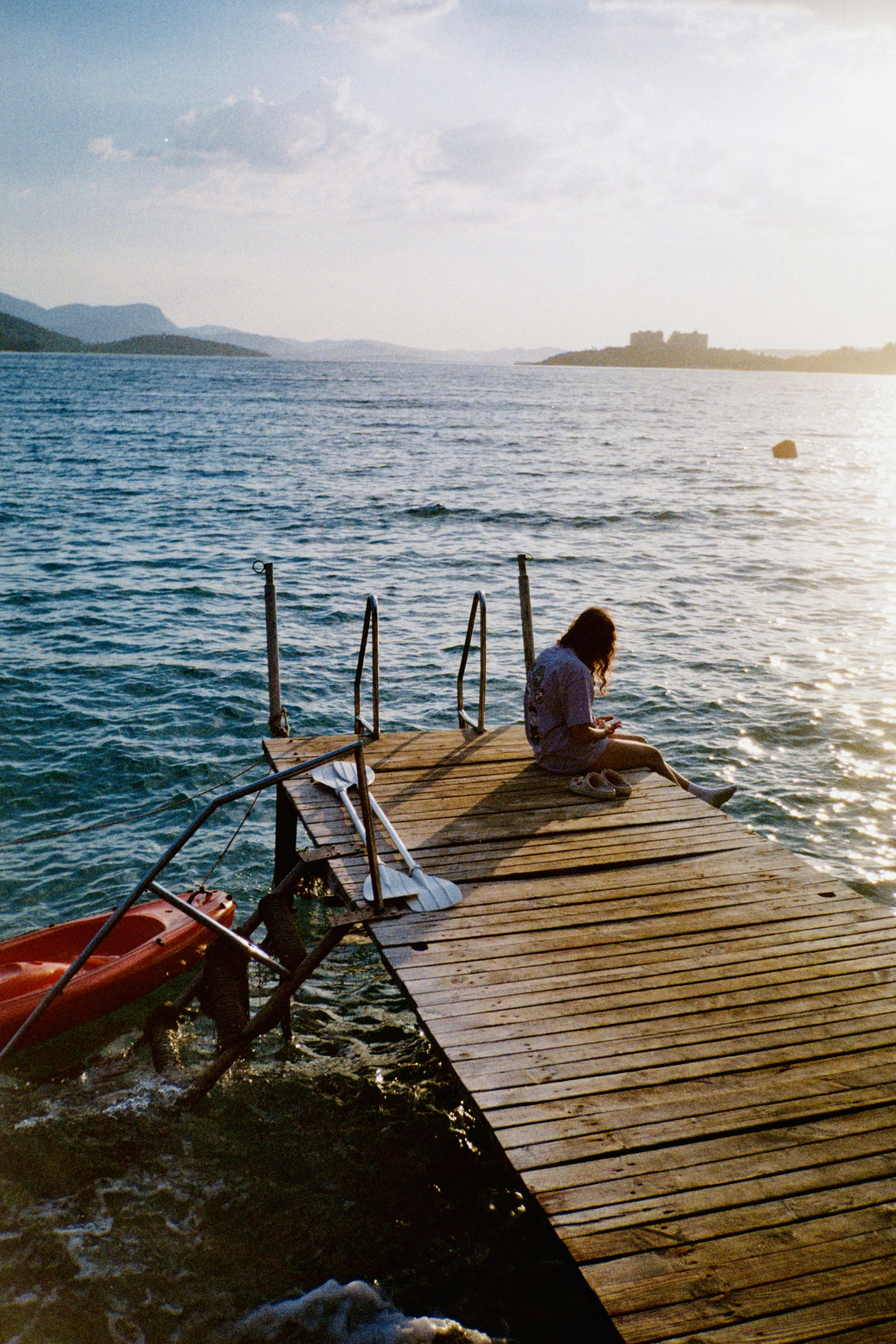Woman sits on wooden pier with red kayak at sunset.