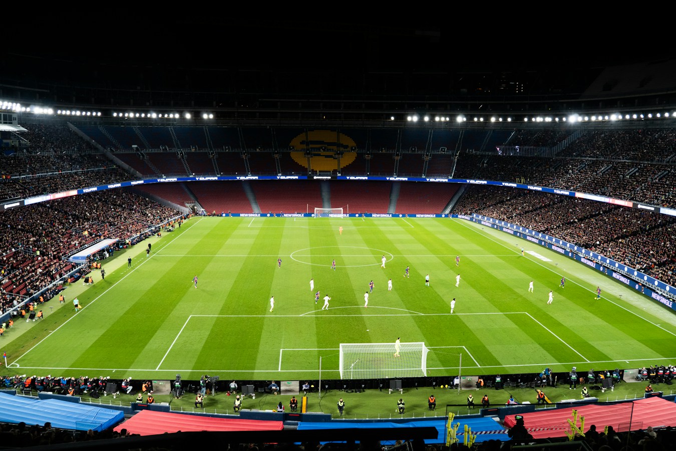 A packed football stadium at night, fully floodlit, with players on a striped green pitch and pitchside advertising boards showing sports-betting