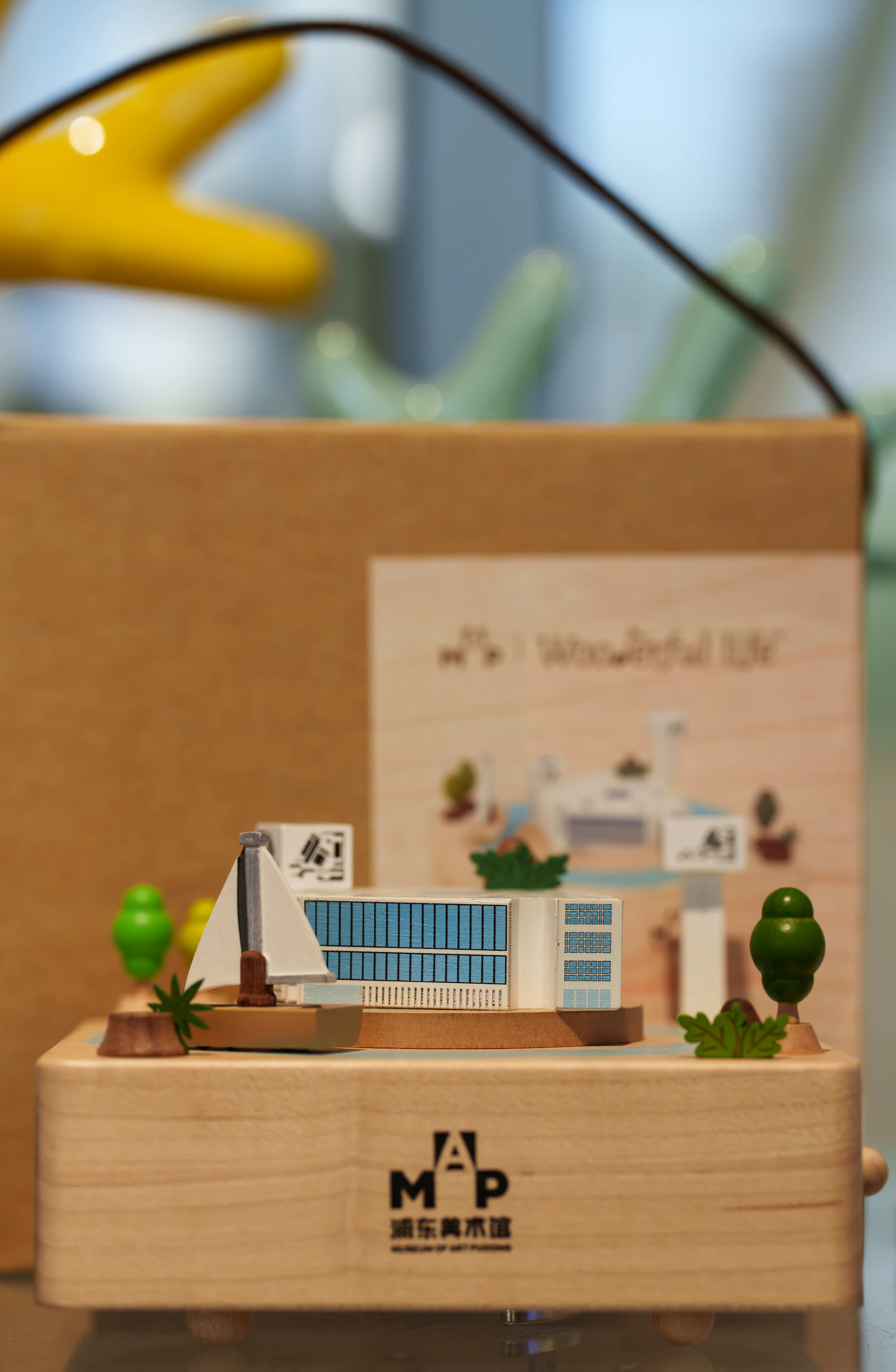 A pale wooden music box with a miniature white building, a sailboat and green trees on its lid, in front of its cardboard packaging