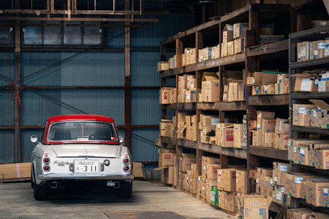 Vintage car parked in a warehouse with shelves of boxes.