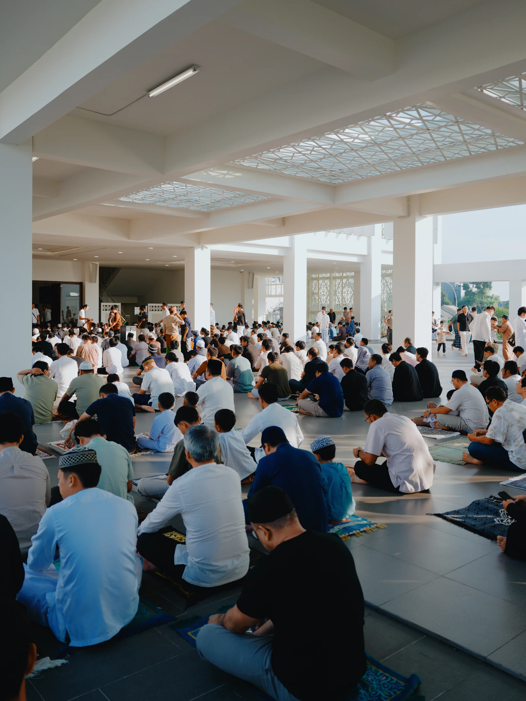 A congregation seated on prayer mats in the open-sided hall of a mosque, waiting for the Eid prayer