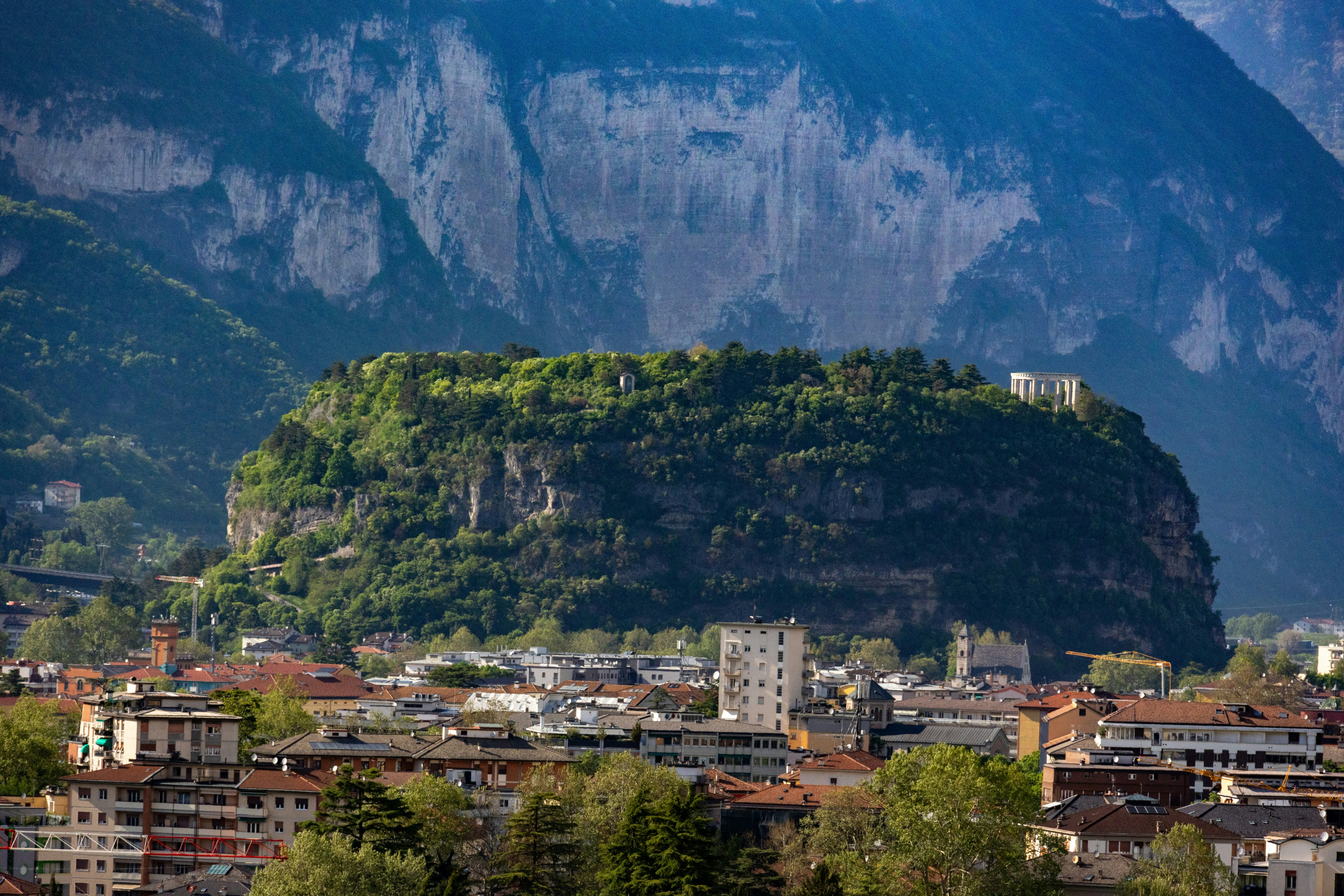 City buildings beneath a tree-covered hill and rugged mountains.