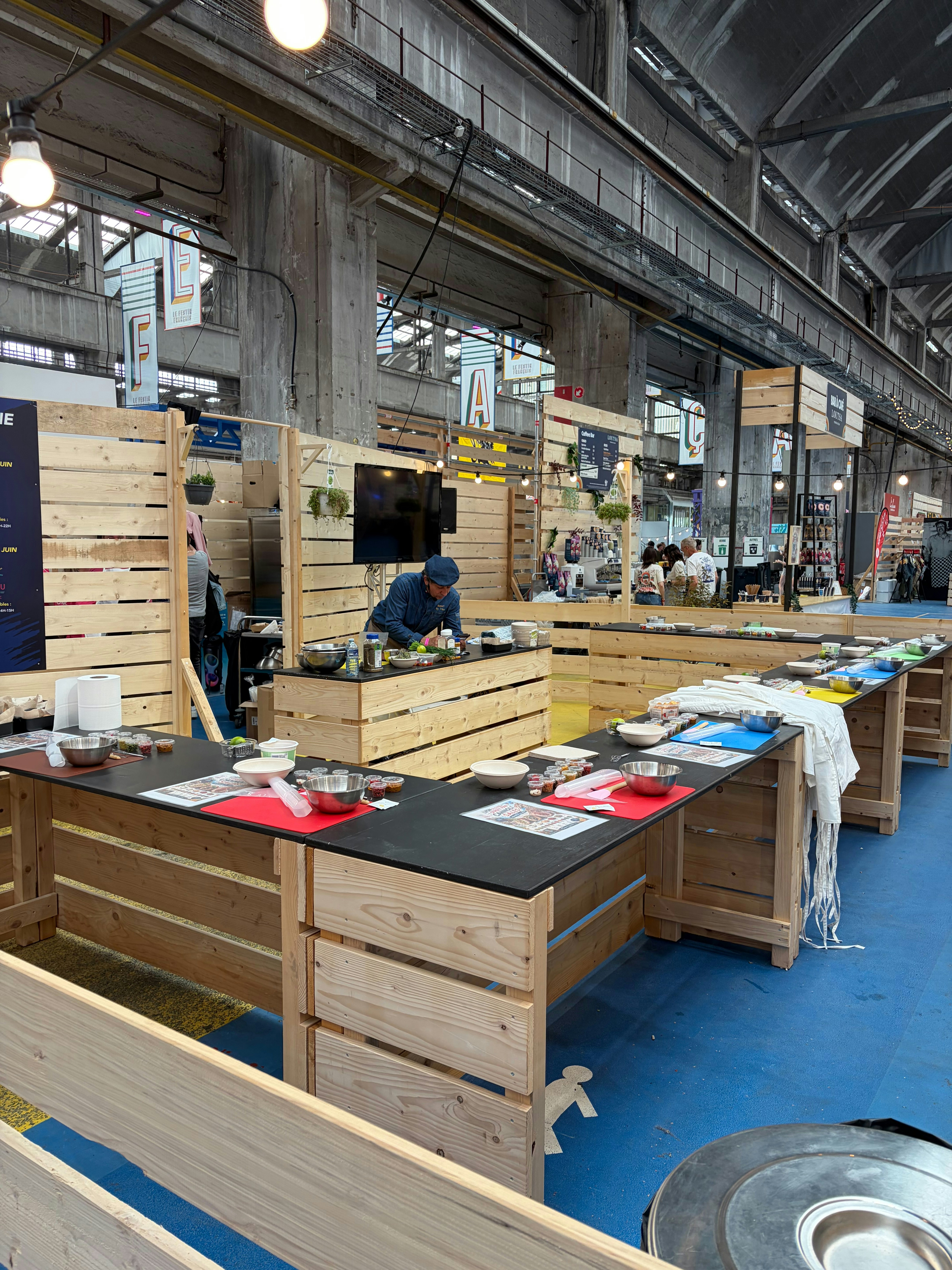 Atelier de démonstration culinaire sur des établis en bois, avec un écran de retransmission, dans la halle du festival