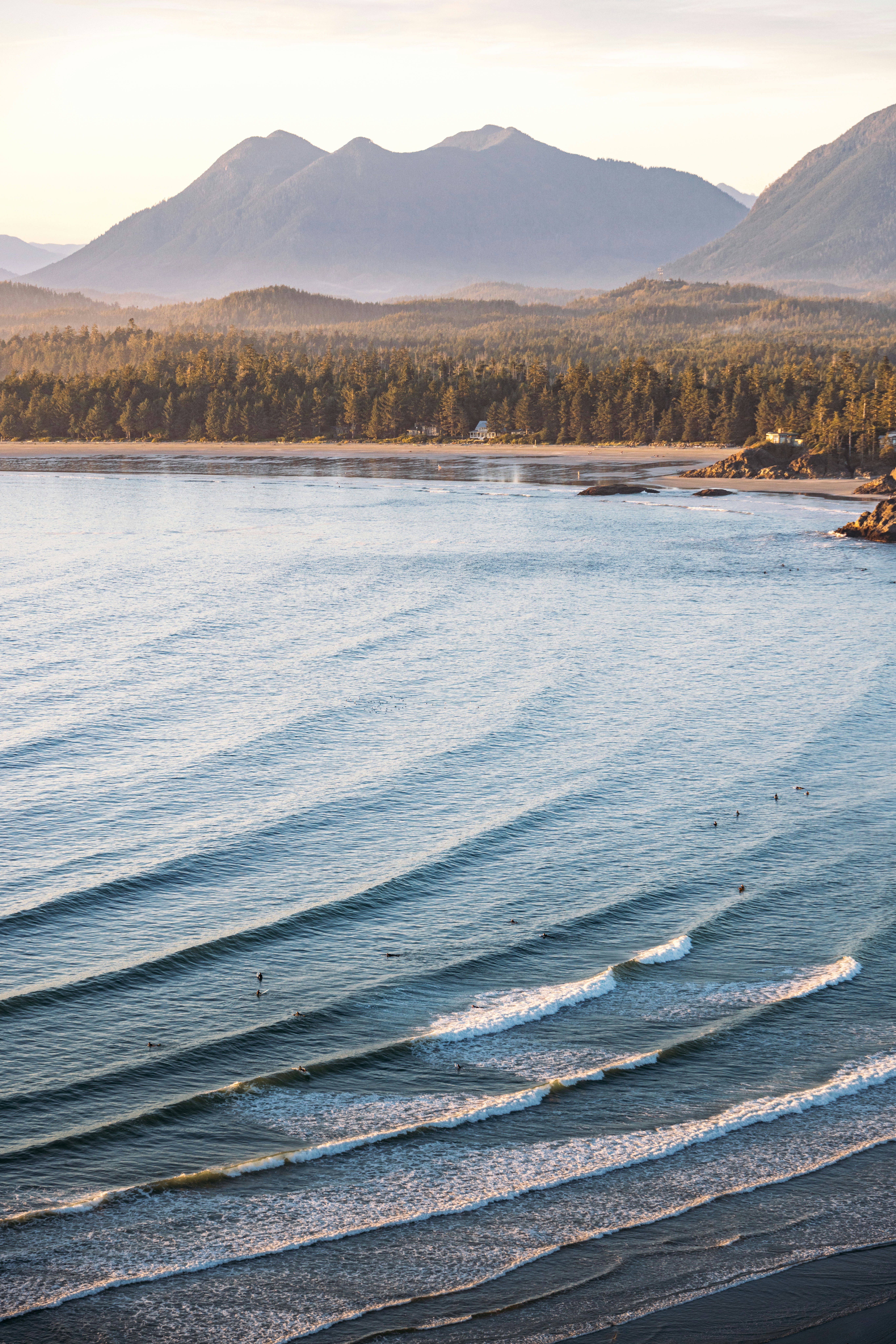 Parallel ocean waves roll towards a forested beach and mountains