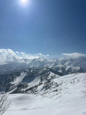 Snow-covered rounded Niigata mountain ridges under a blue sky, with a groomed piste in the foreground