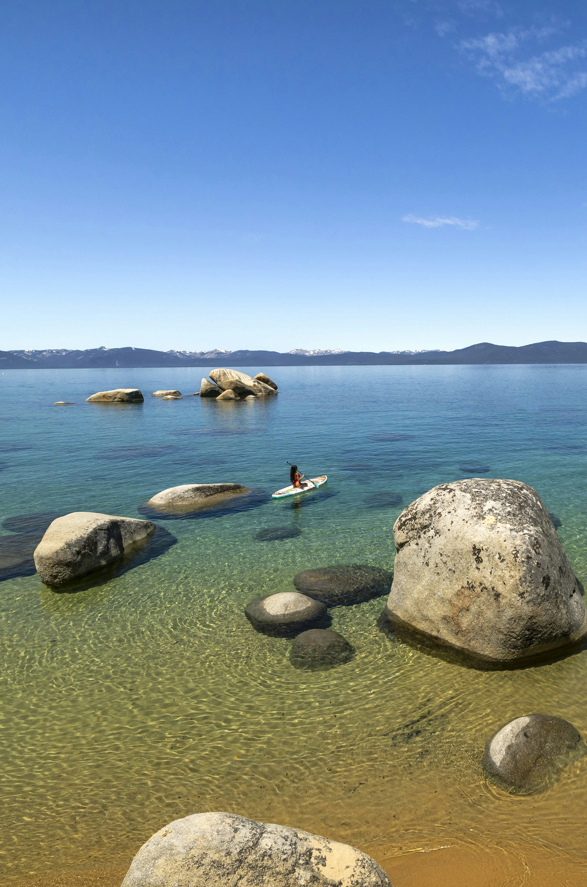 Paddleboarder on crystal clear lake surrounded by large boulders.