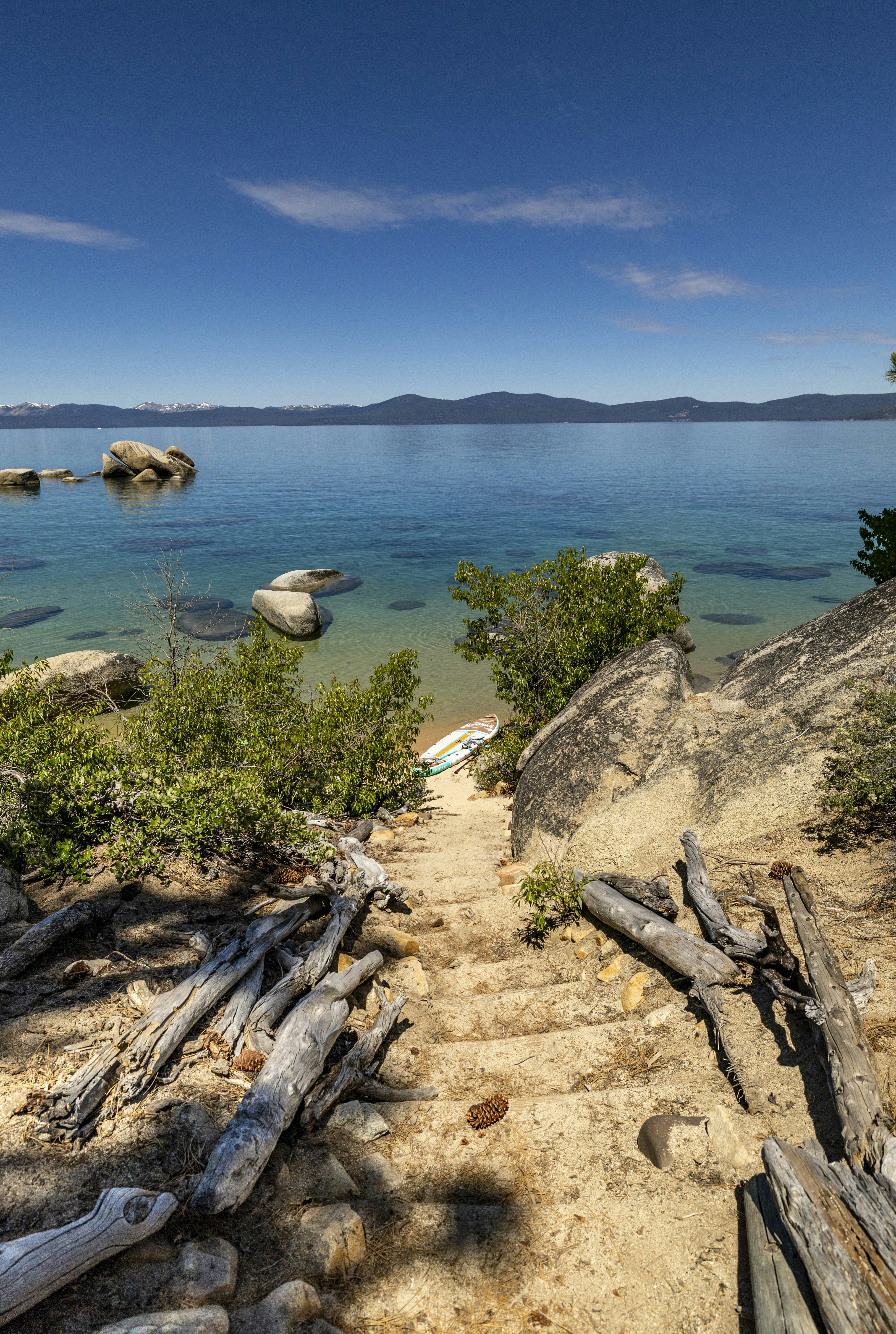 Clear blue lake, sandy path, driftwood, and distant mountains.