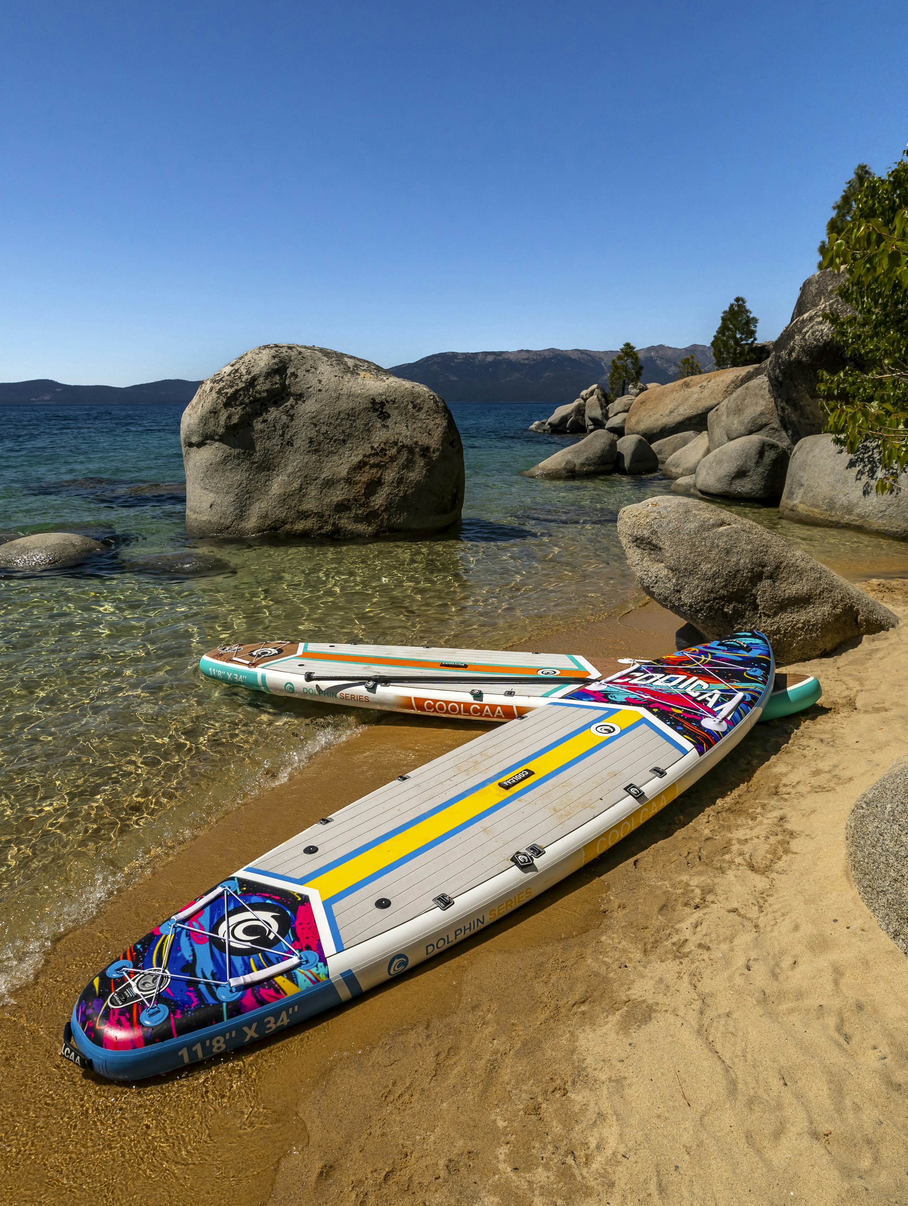 Colorful paddleboards on a sandy beach by clear lake water.