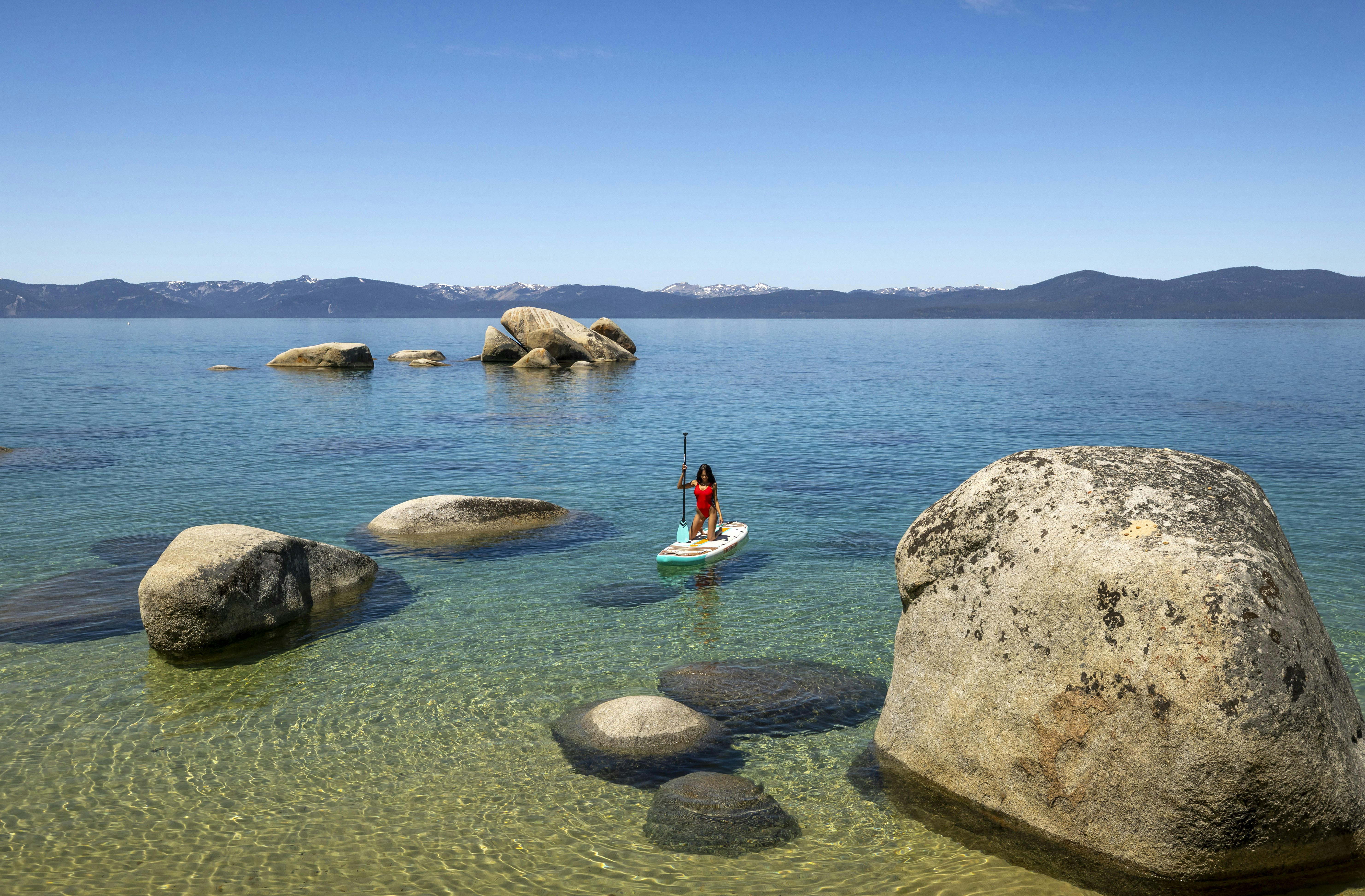 A person paddleboarding on clear blue water with large rocks.