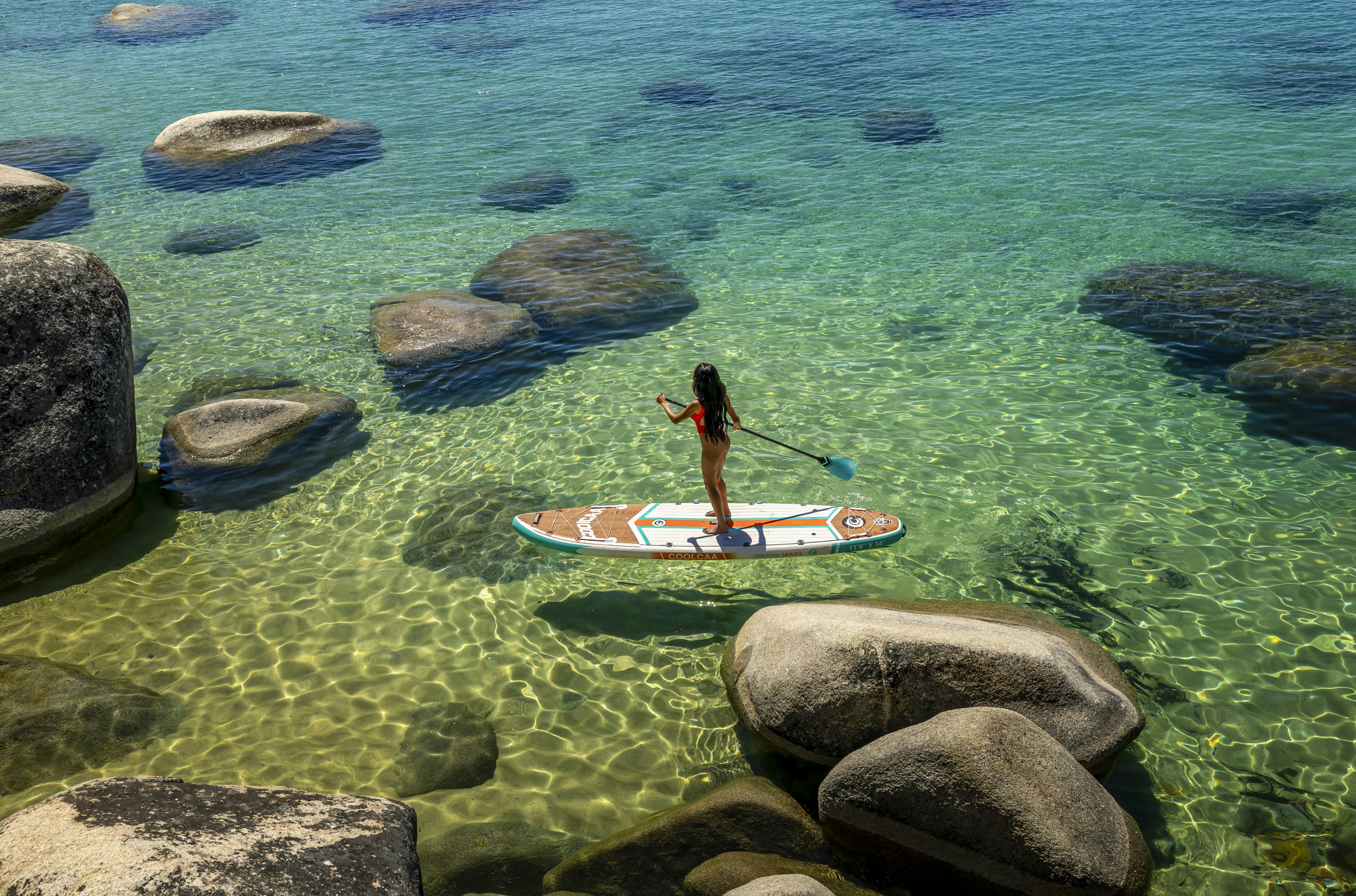 A woman paddleboarding on clear water with large rocks.