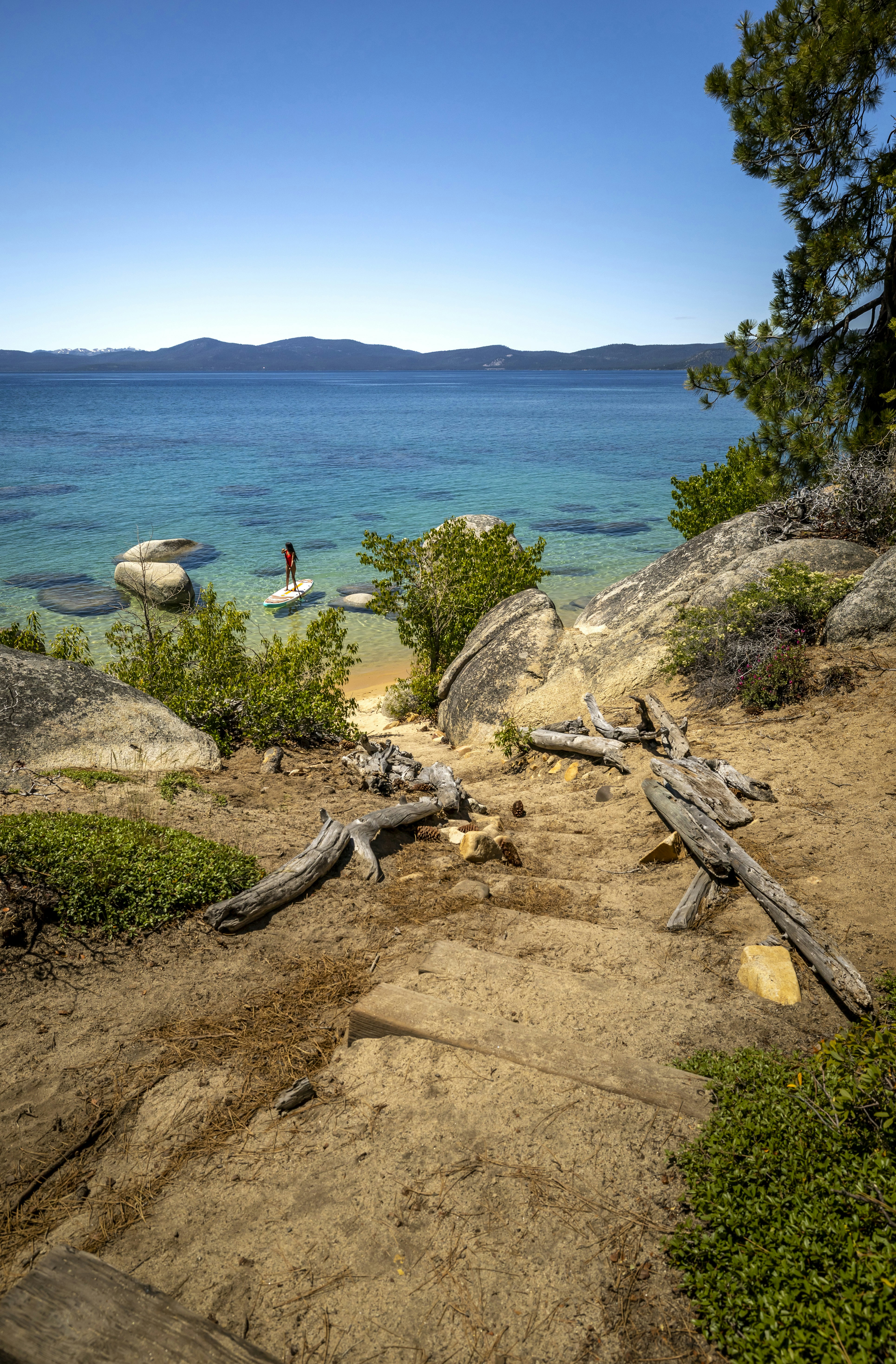 Serene blue lake with a paddleboarder and rocky, sandy shore.