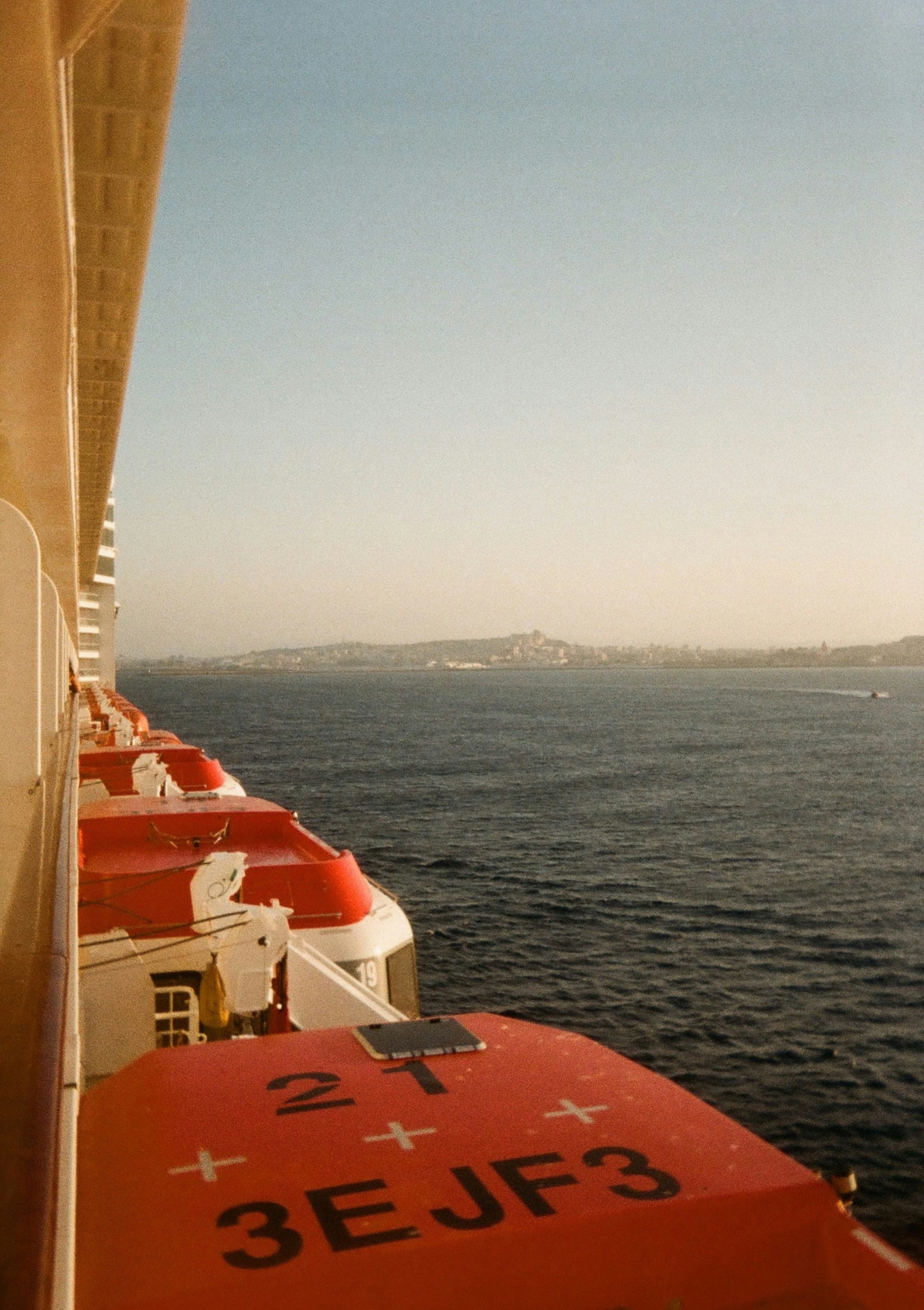 Ship's side with red lifeboats, blue sea, and distant island.