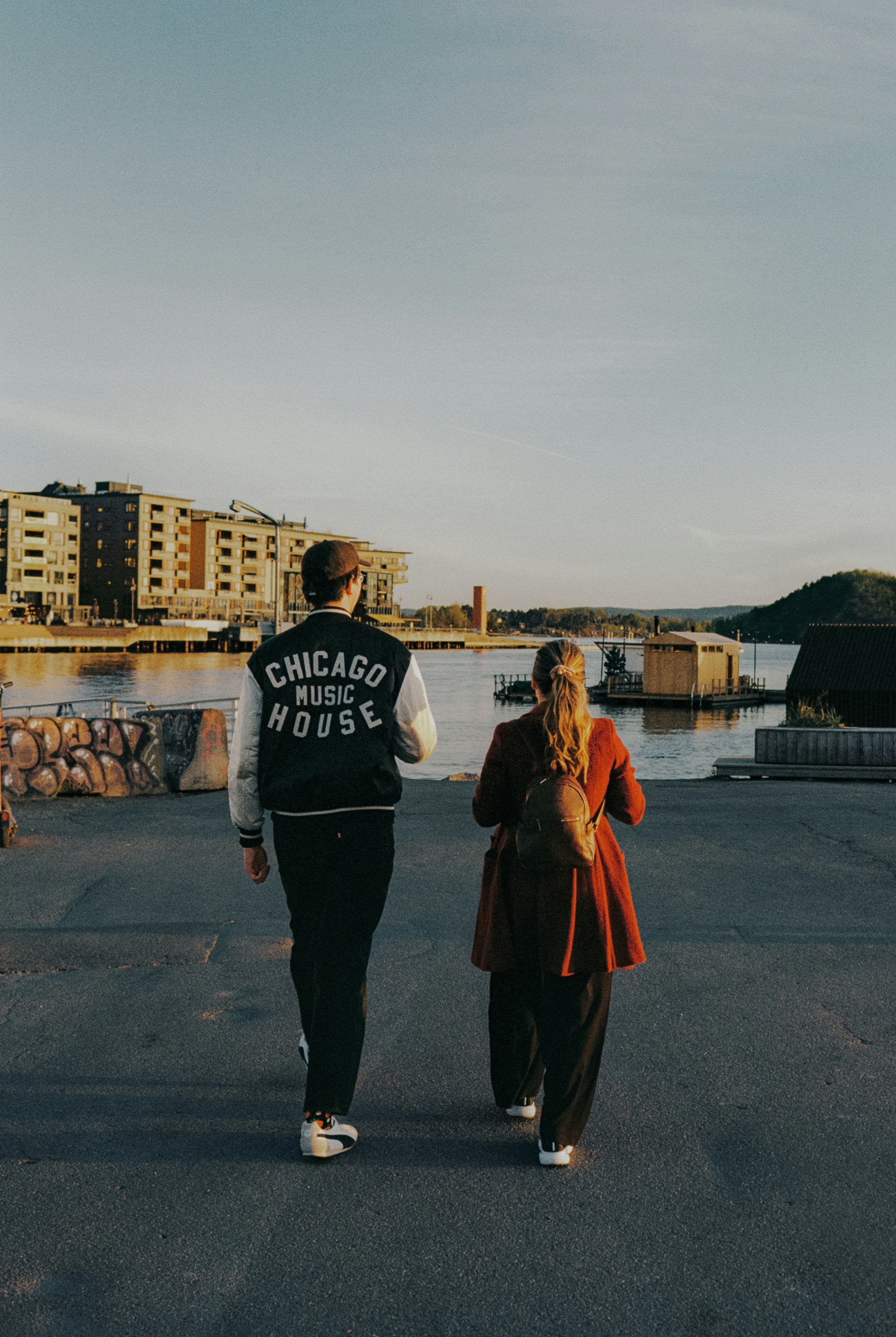 Two people walk towards buildings by the water at sunset
