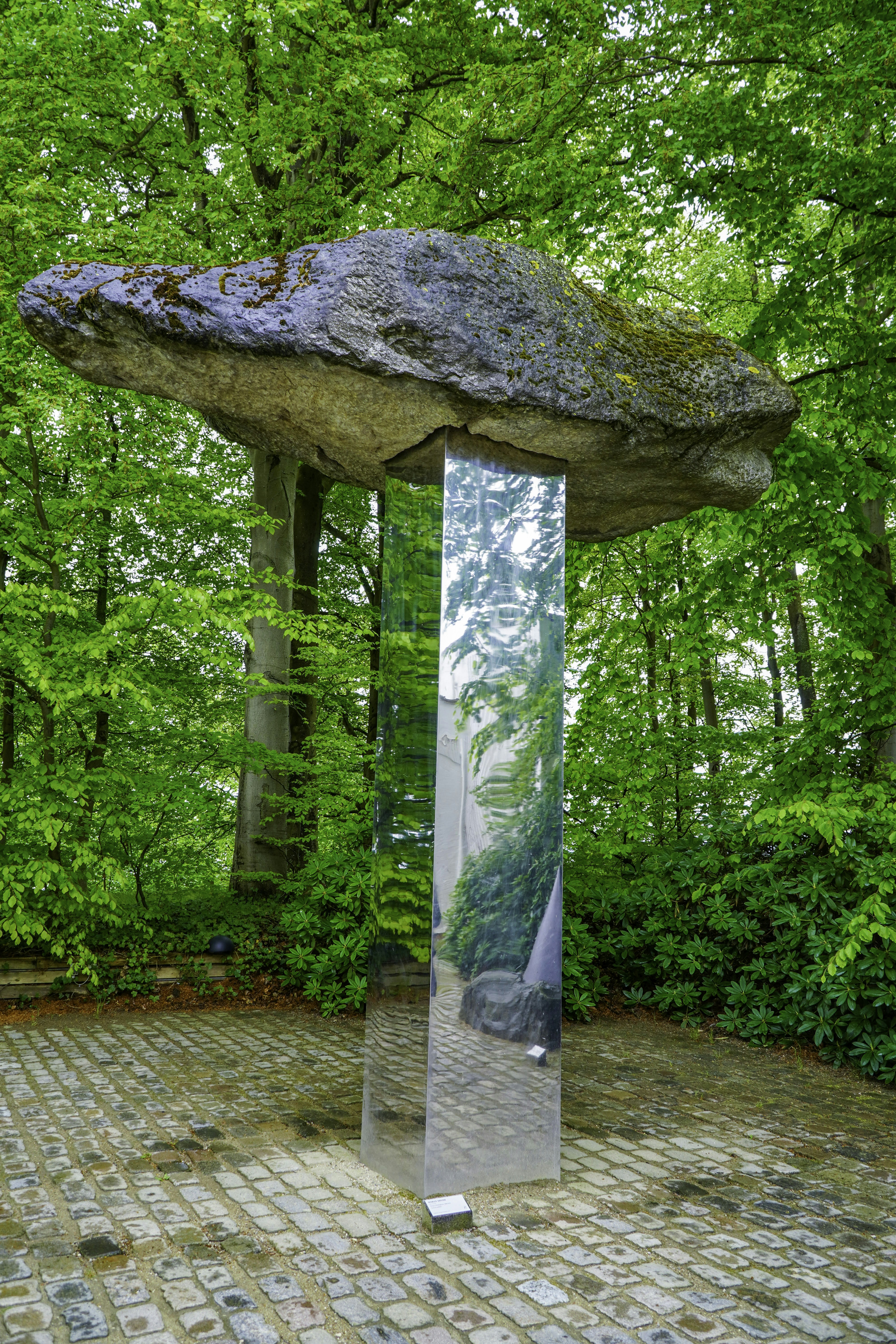 Boulder balanced on a mirrored column amidst vibrant green trees.
