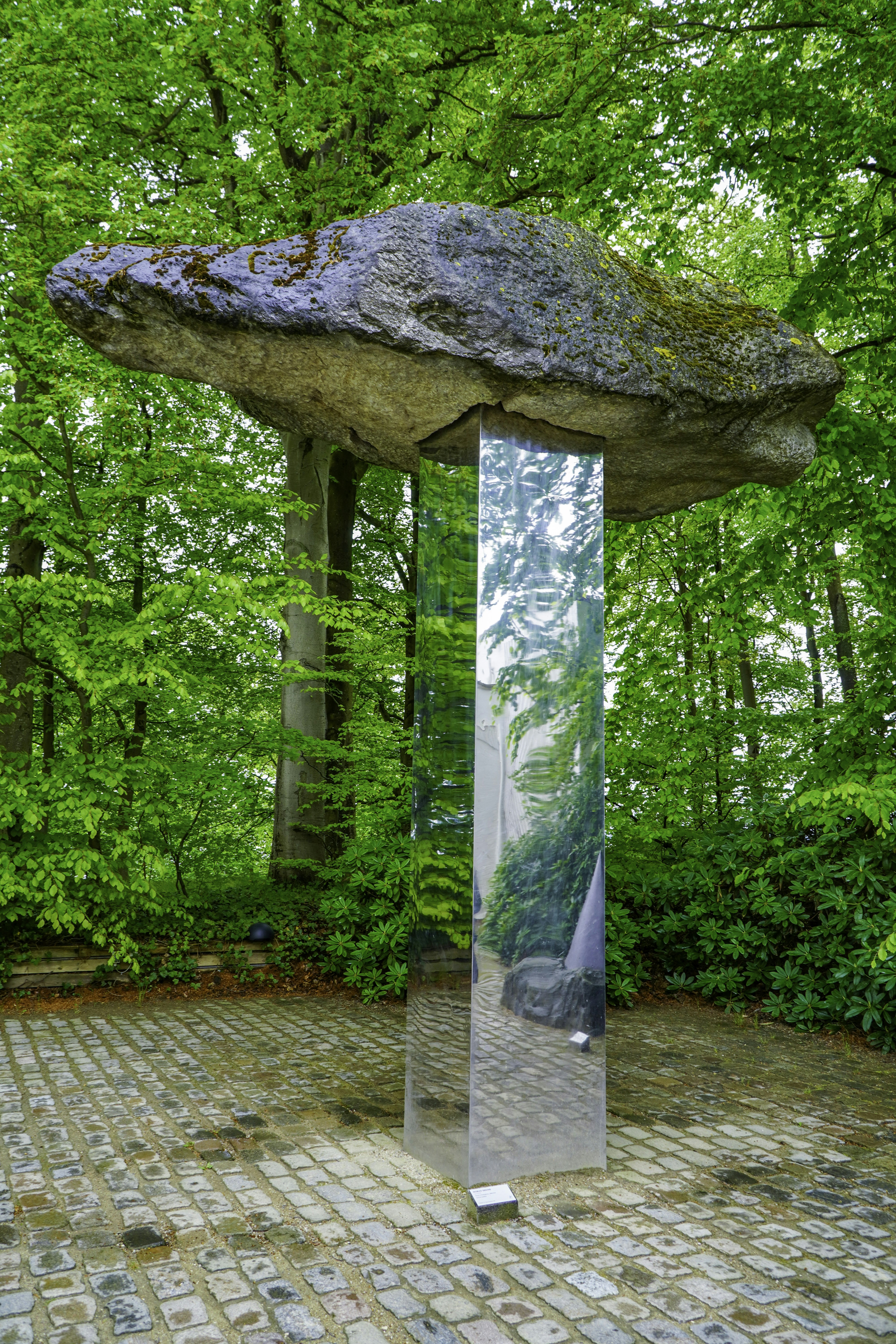Stone boulder balanced on a reflective column amidst green trees.