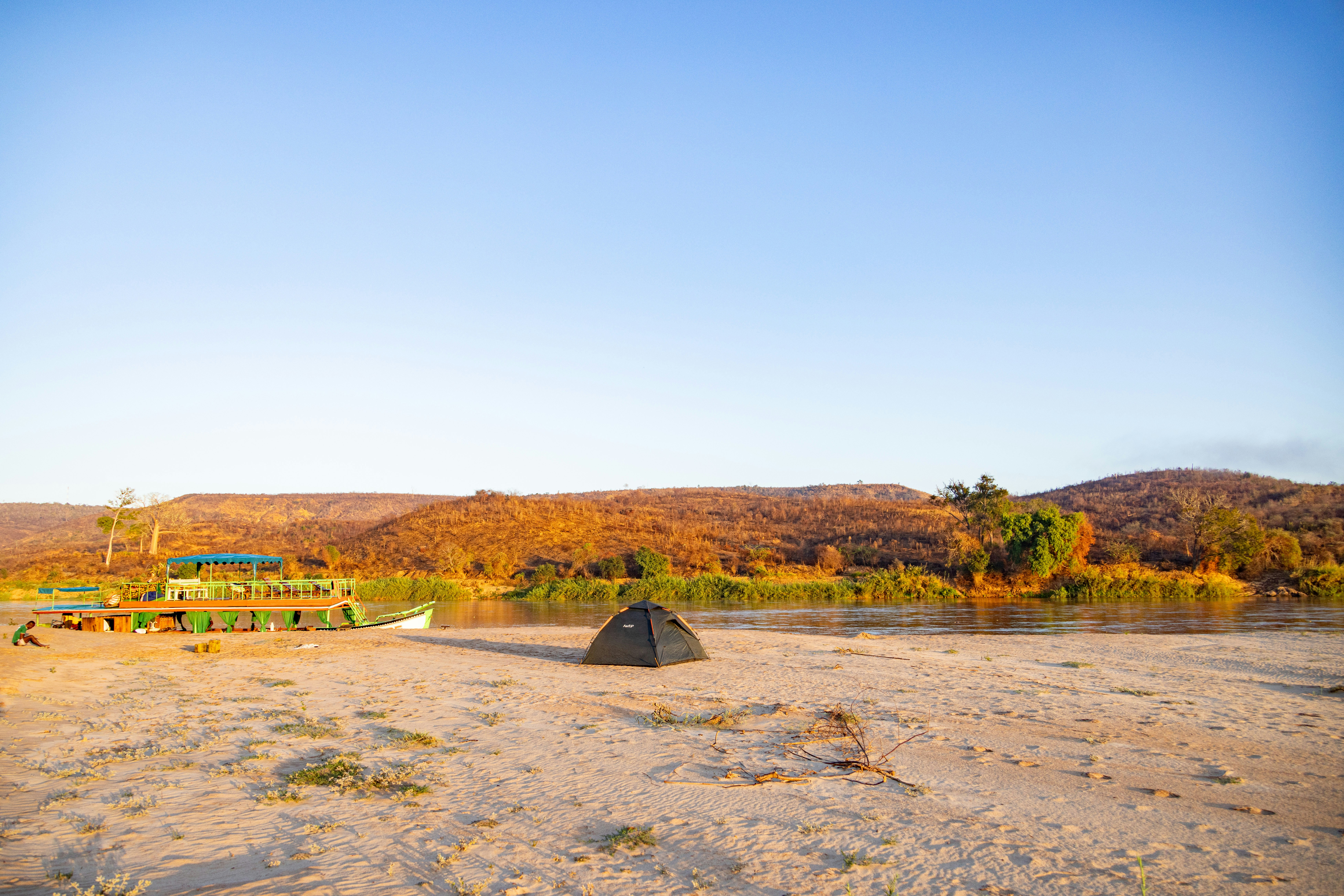 Tienda negra en una playa de arena junto al río con colinas otoñales.