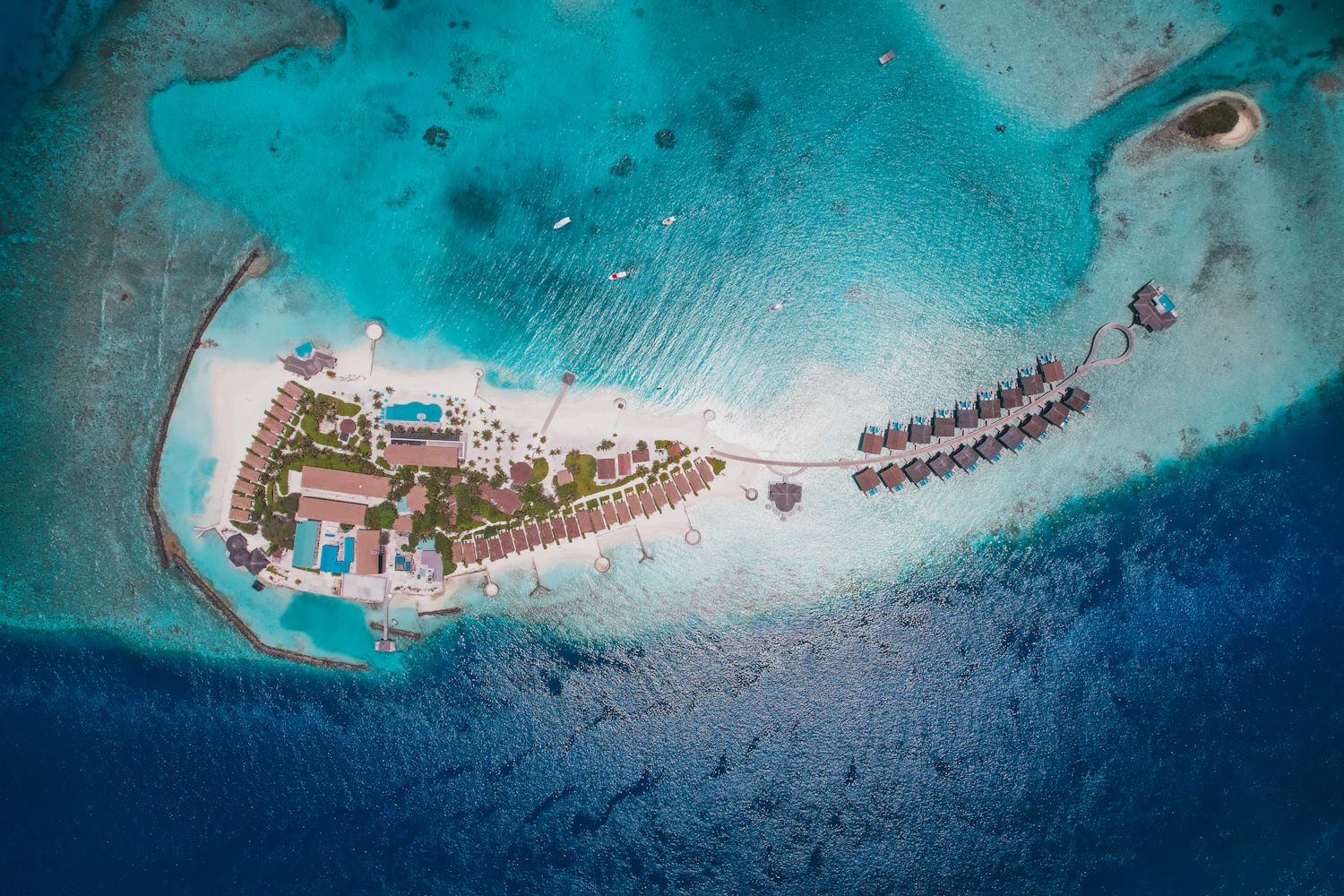Aerial view of a small tropical island resort with a curve of overwater villas in a turquoise lagoon.