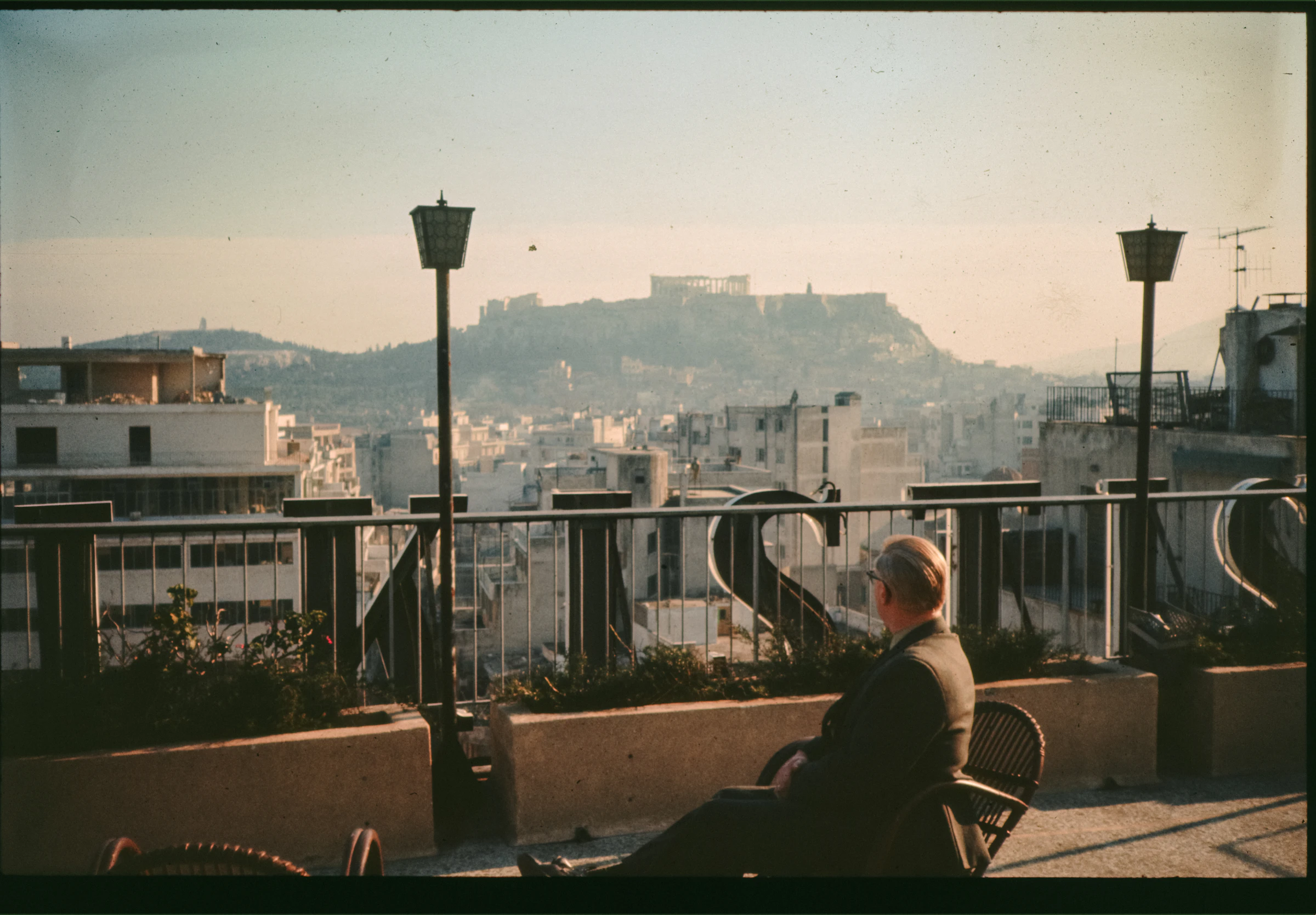 A man sits alone on a sunlit rooftop terrace looking out over the city