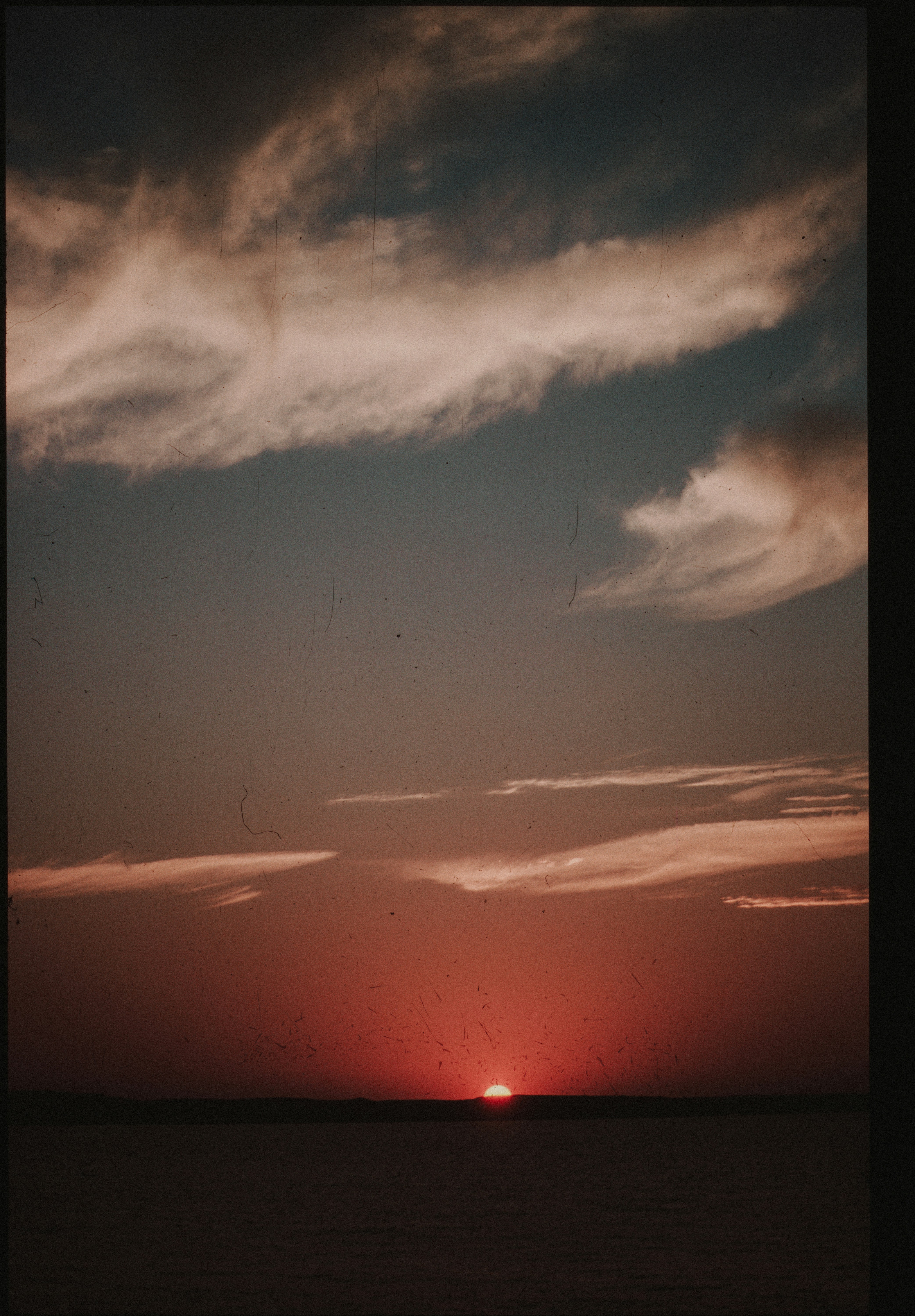 Vibrant red sunset over calm water with wispy clouds.