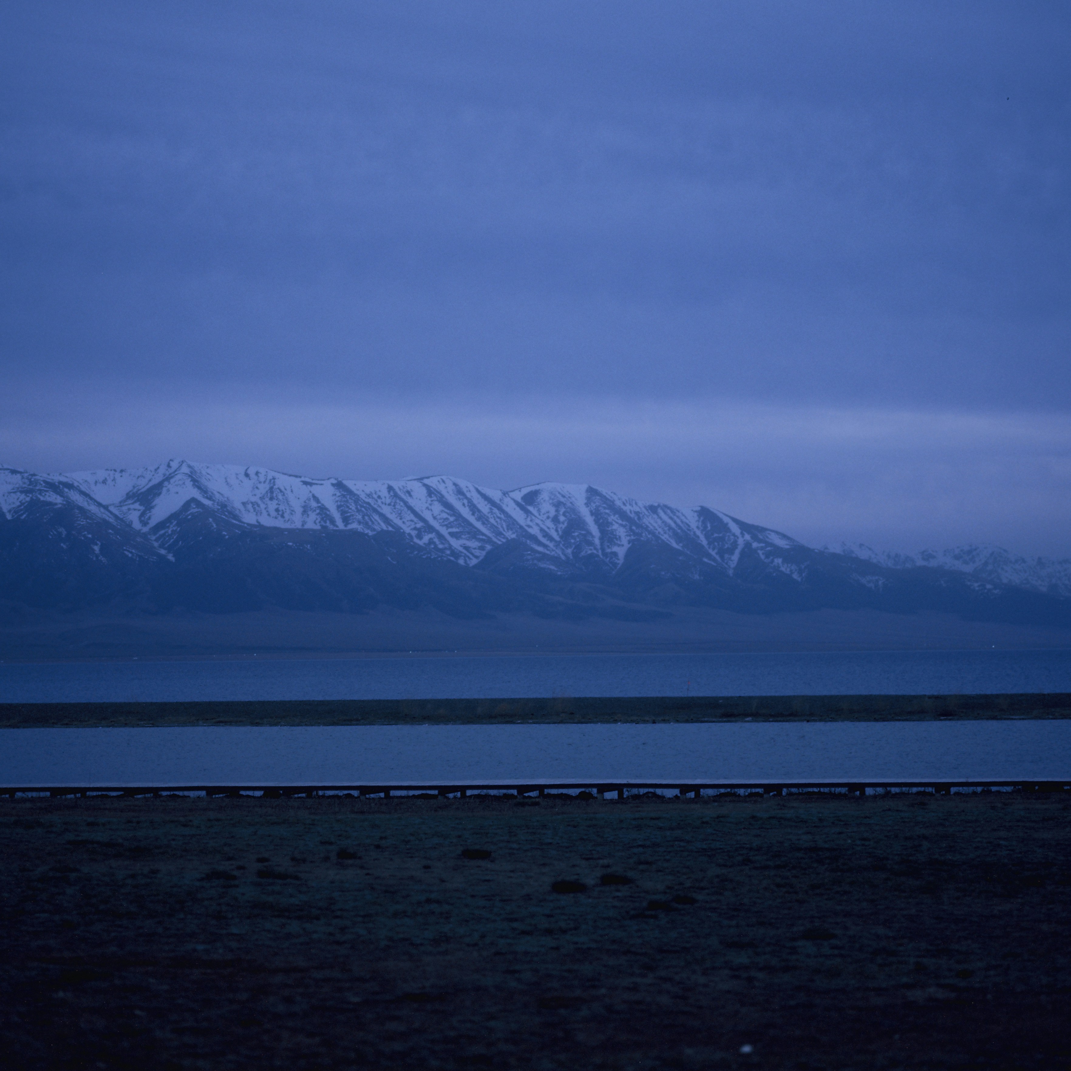 Snow-capped mountains overlook a calm lake under a dark sky.