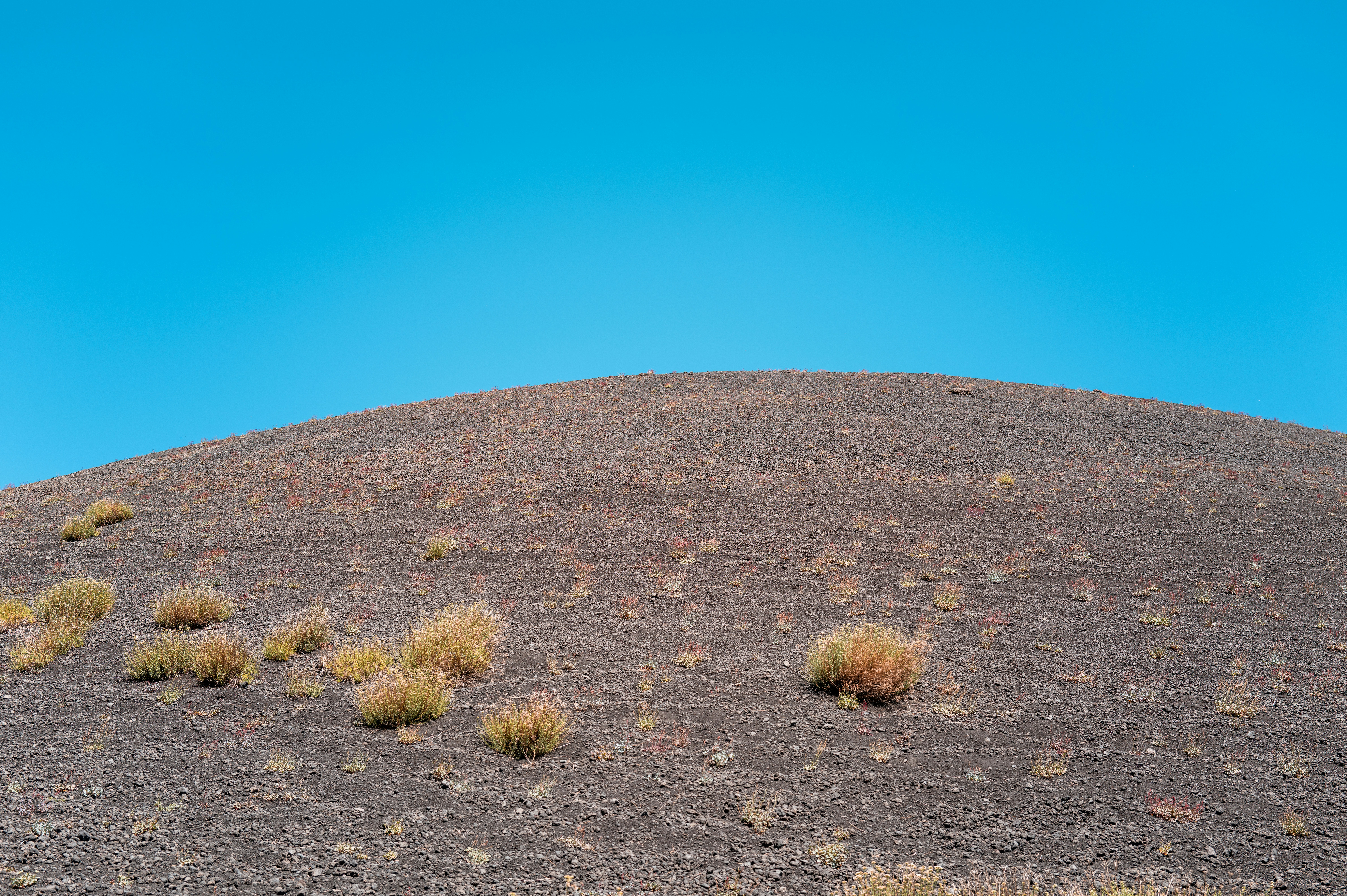 Barren rocky hill with sparse dry plants under blue sky.