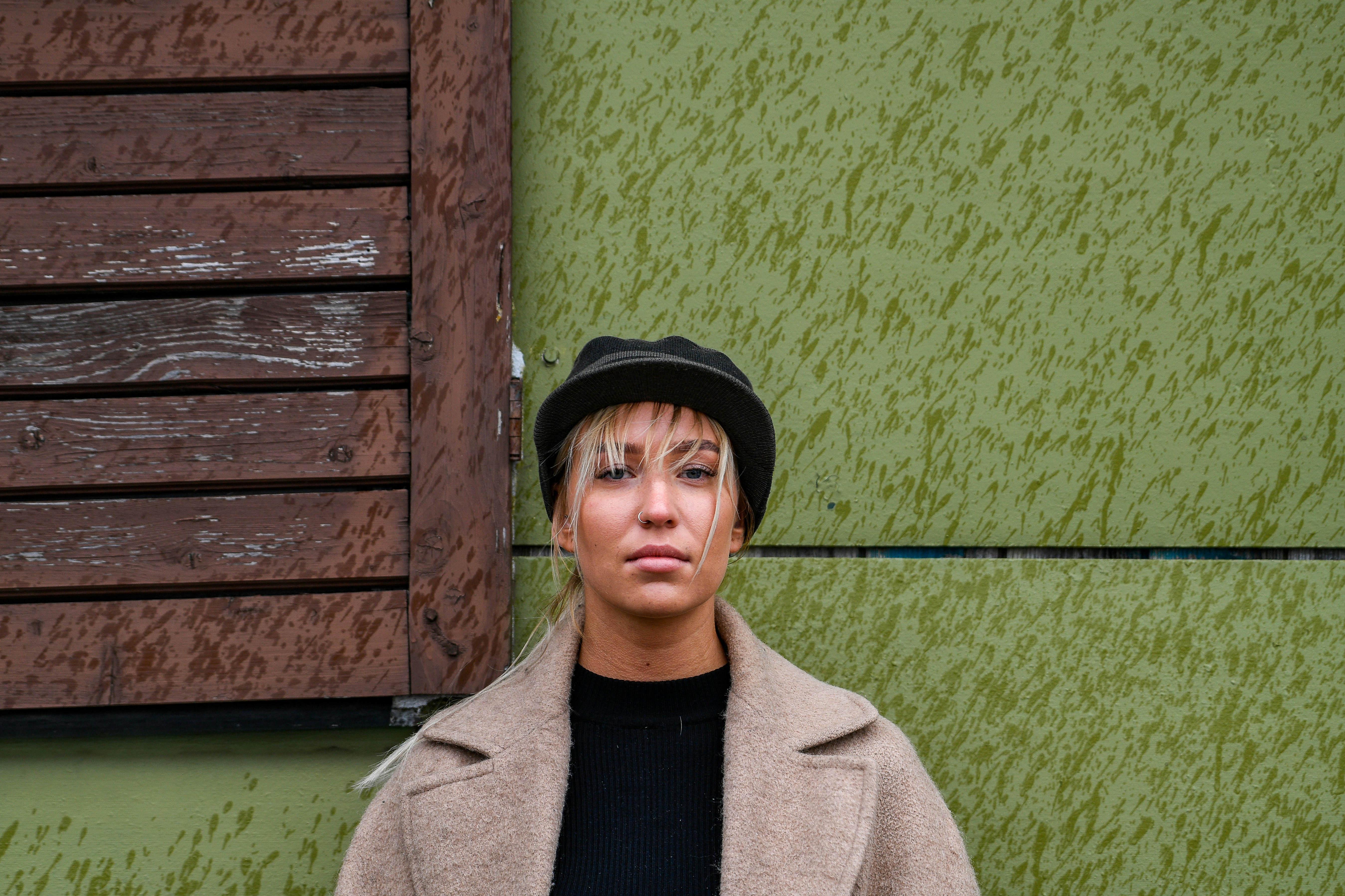 Donna bionda con cappello e cappotto contro una parete verde testurizzata.