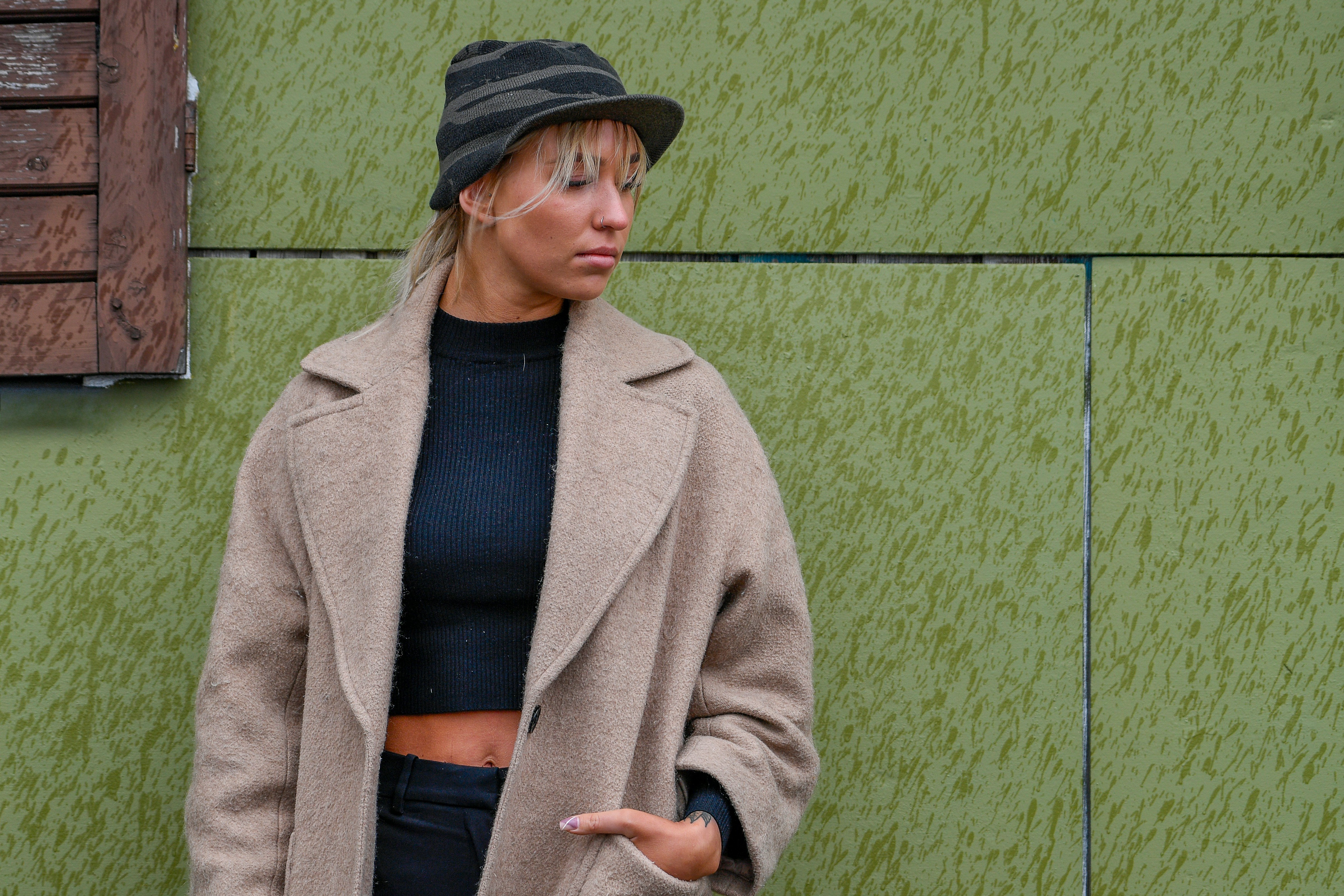Donna con cappotto beige e cappello a righe contro il muro verde.