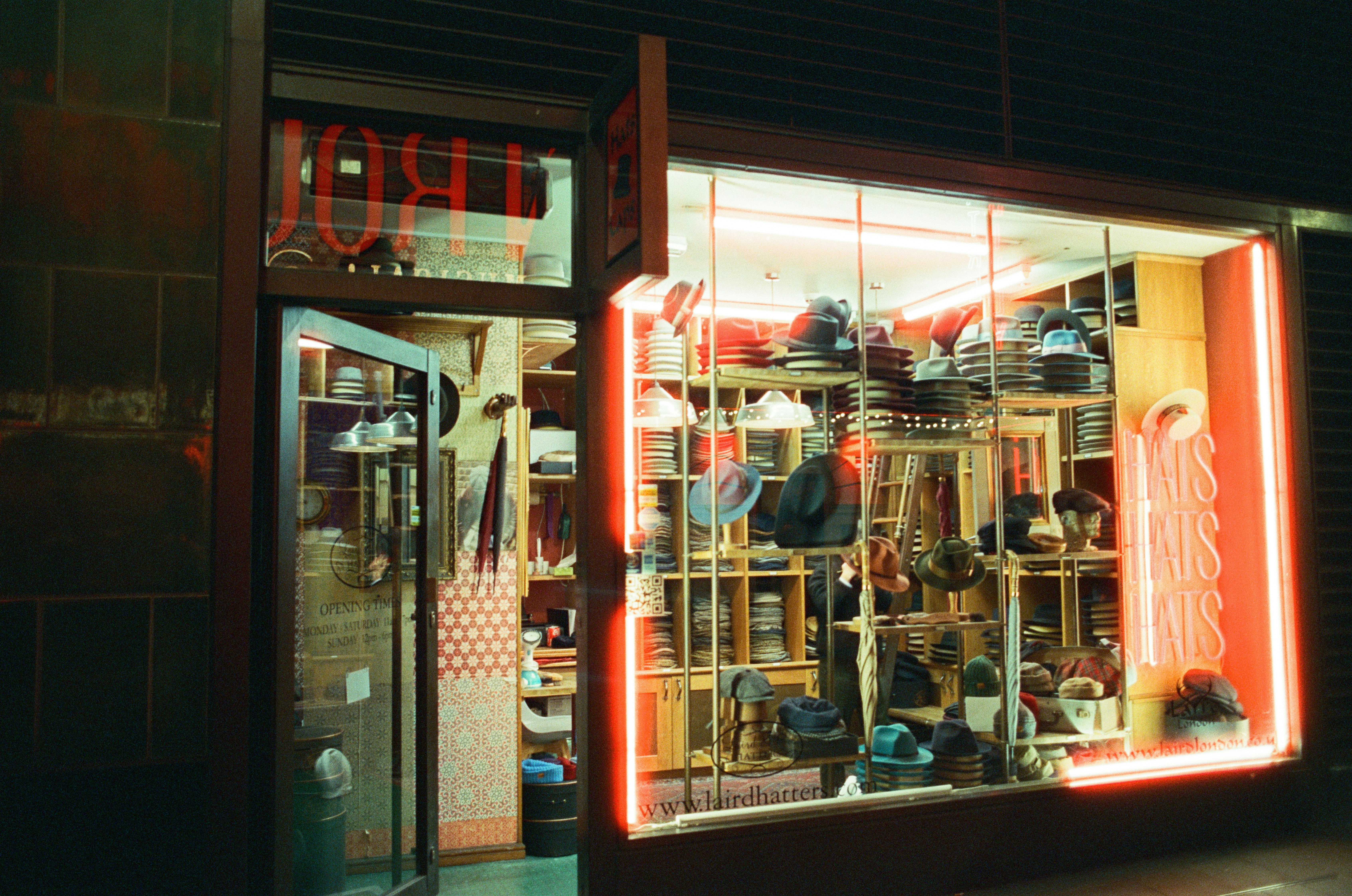A glowing shop window crammed with hats lights up a dark street at night.