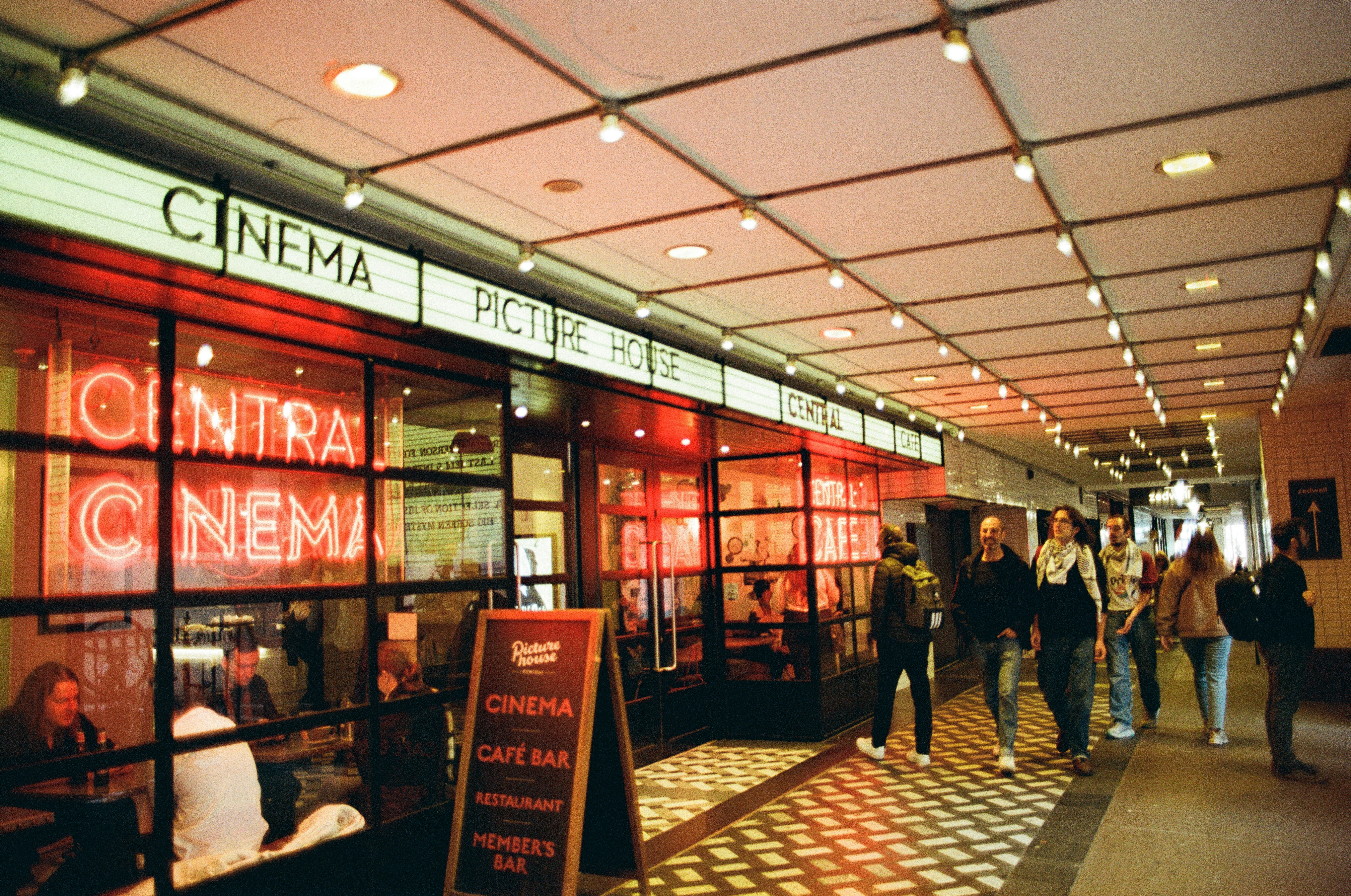 Red neon cinema signs glow inside an arcade as people walk past a Picturehouse cafe bar.