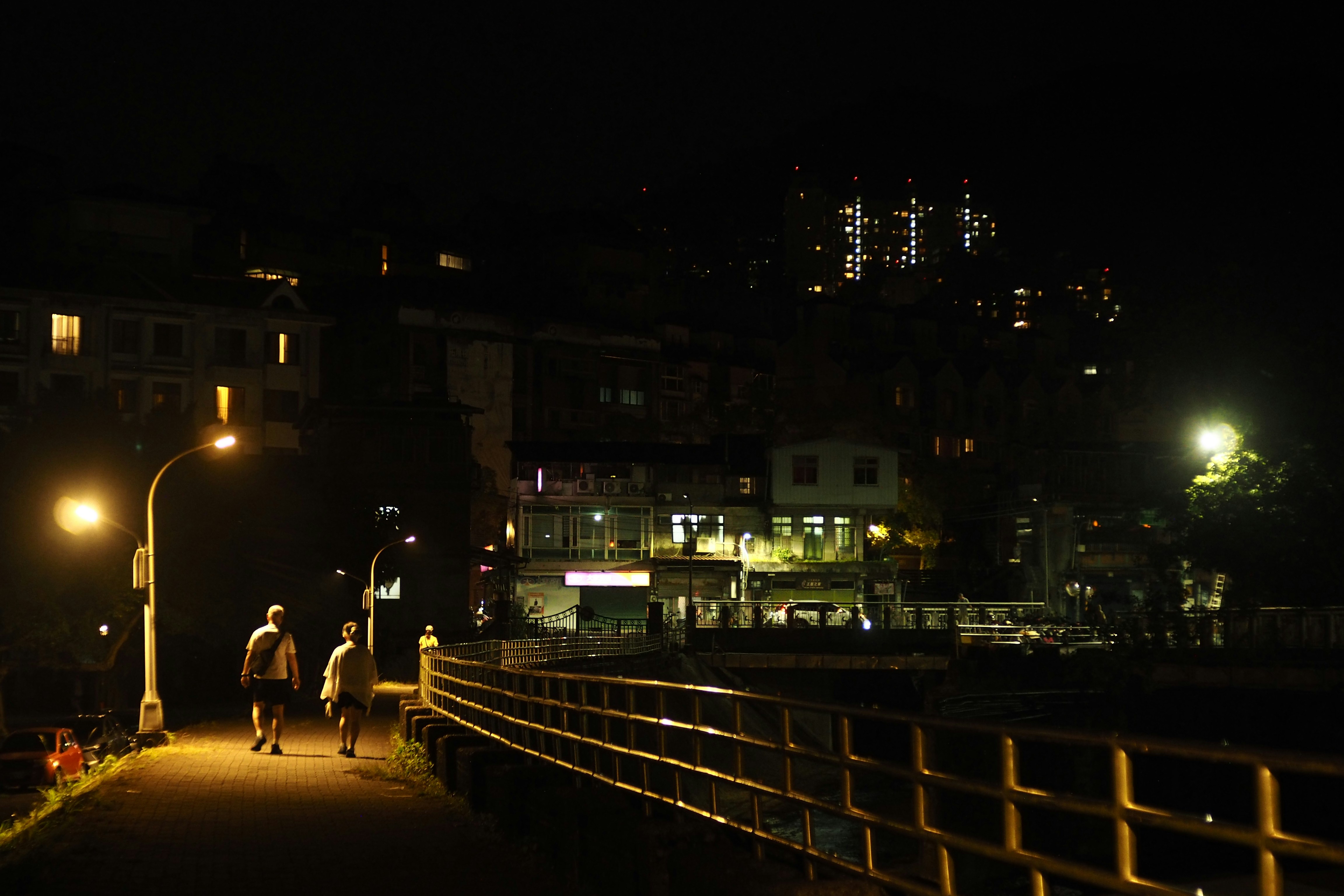 Two people walk past a streetlamp at night with buildings.
