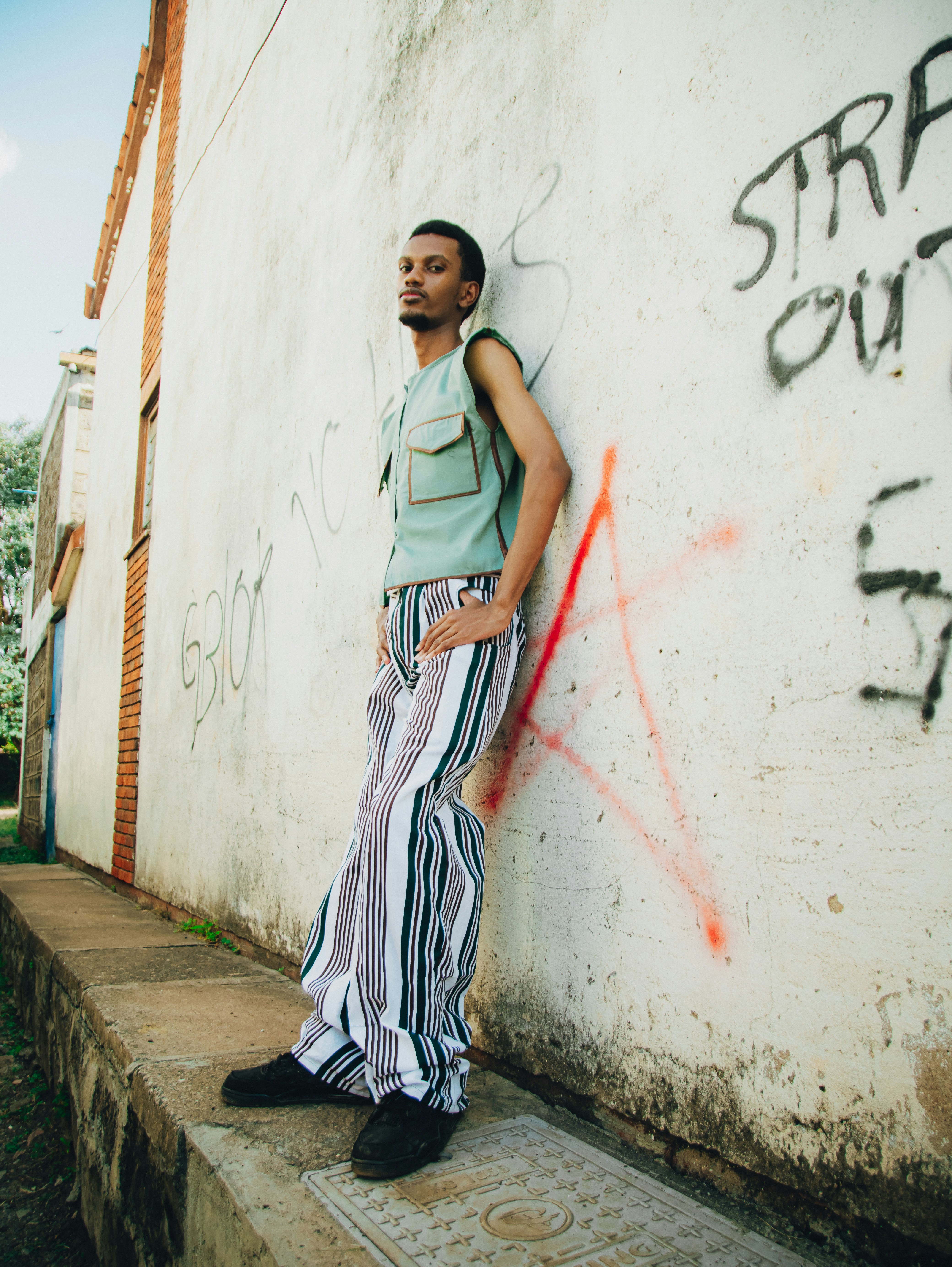 A man in striped pants leaning against a graffiti-covered wall.