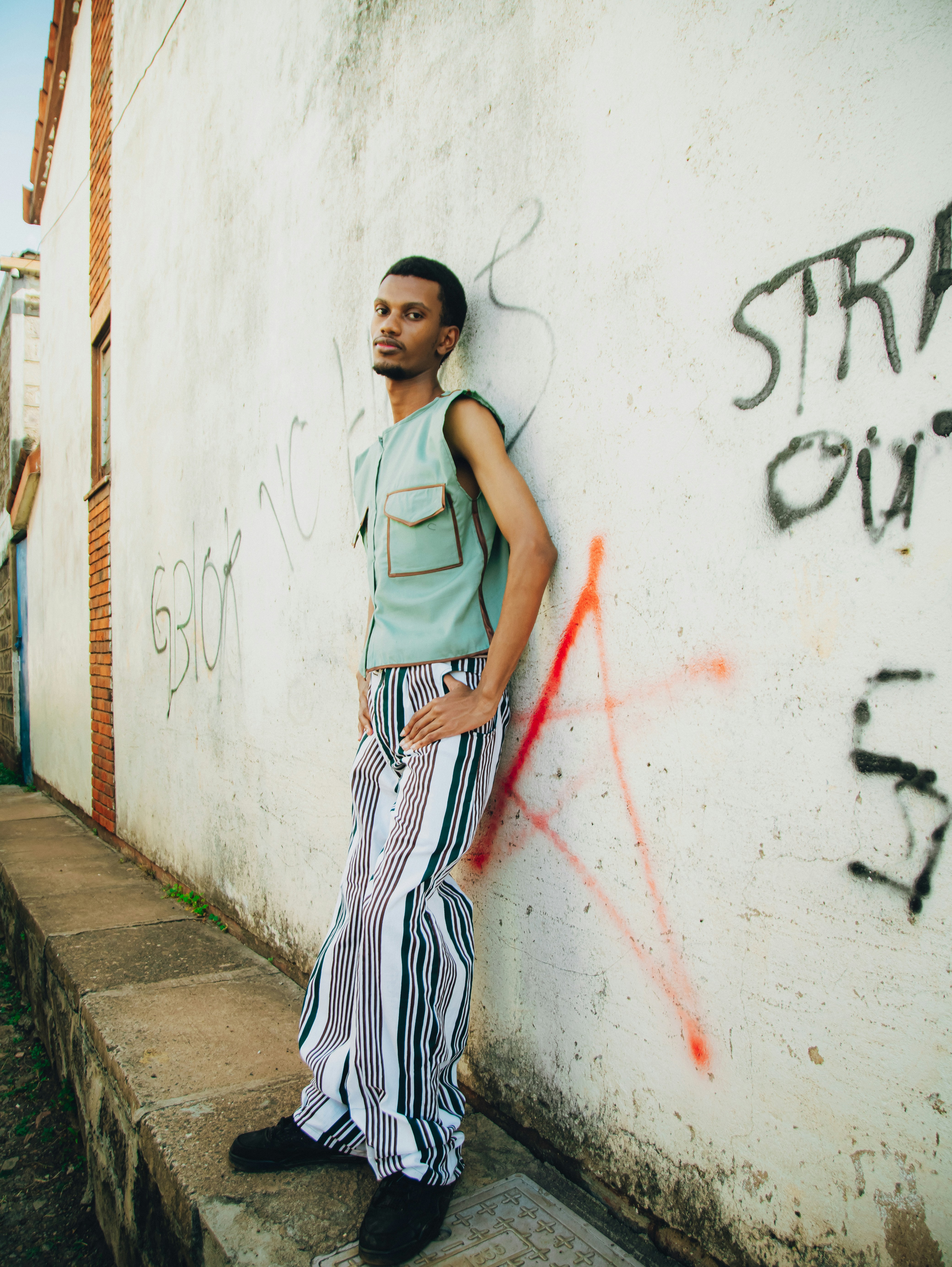 Man in green vest and striped pants against graffiti wall.