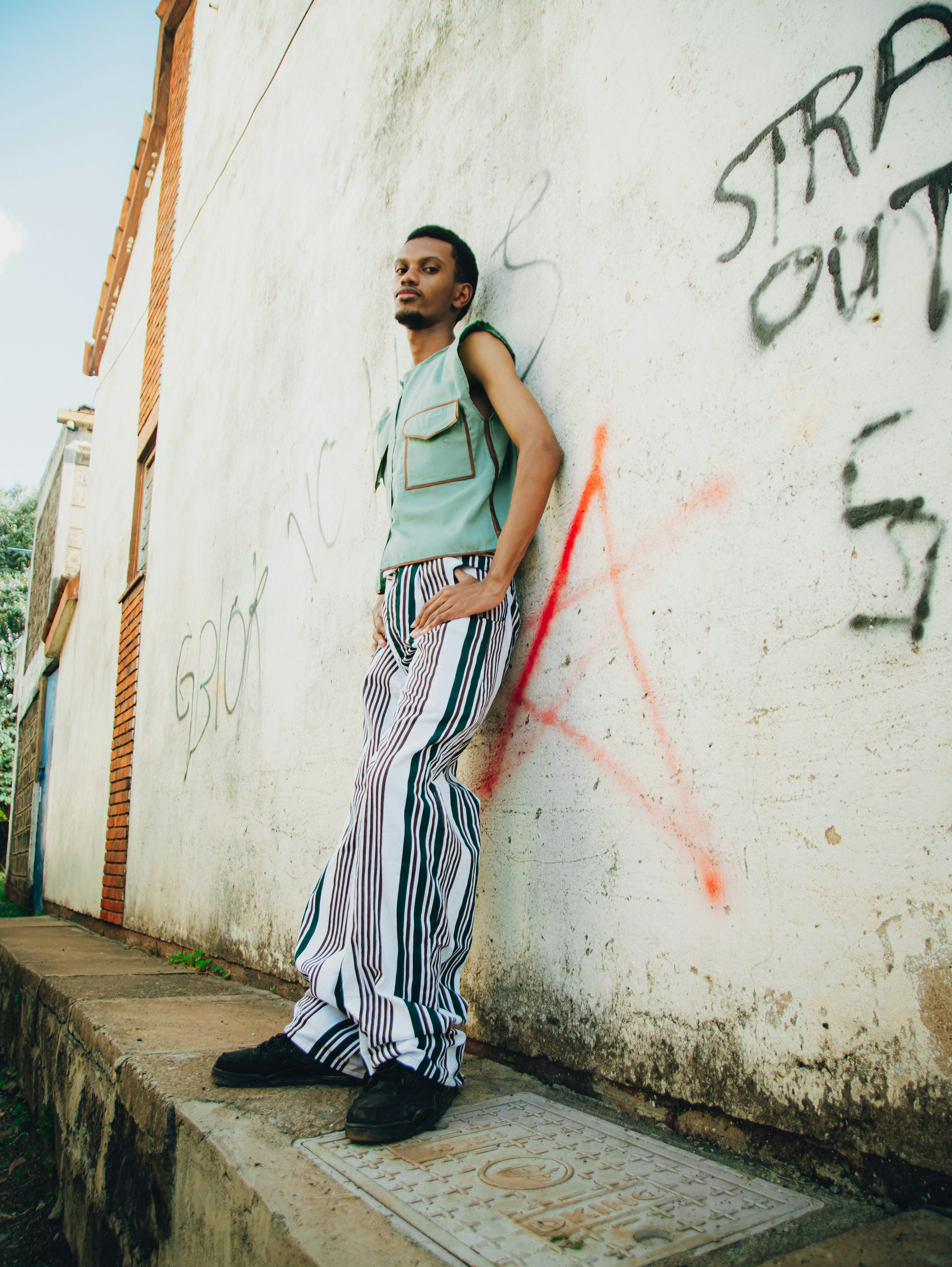A man in striped pants leans against a graffiti-covered wall.