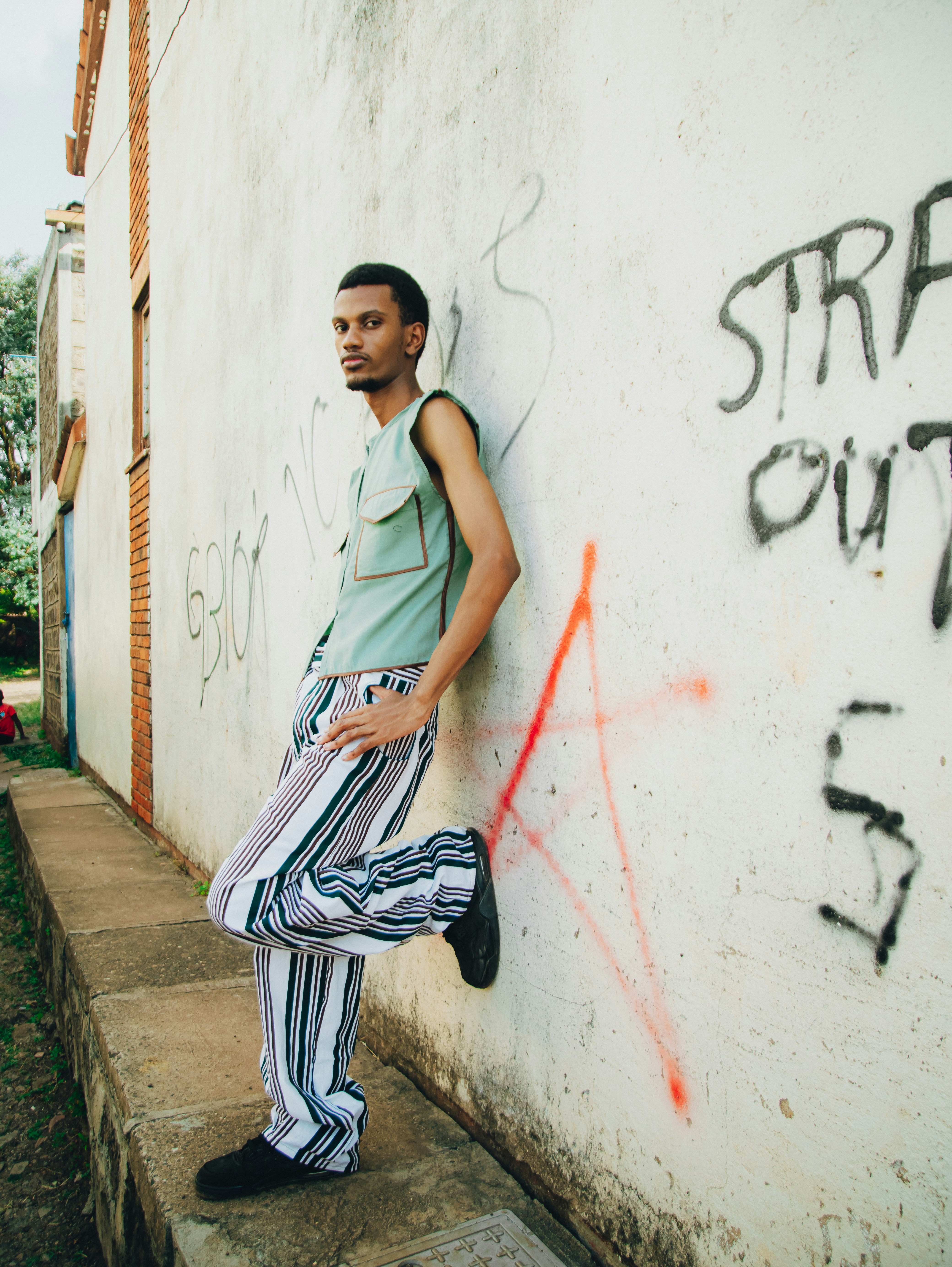 A man in striped pants leans against a graffiti wall.